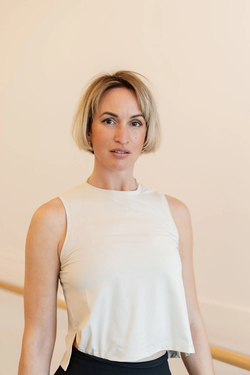 A woman with short blonde hair wearing a sleeveless off-white top and black pants standing against a plain, light-colored background.