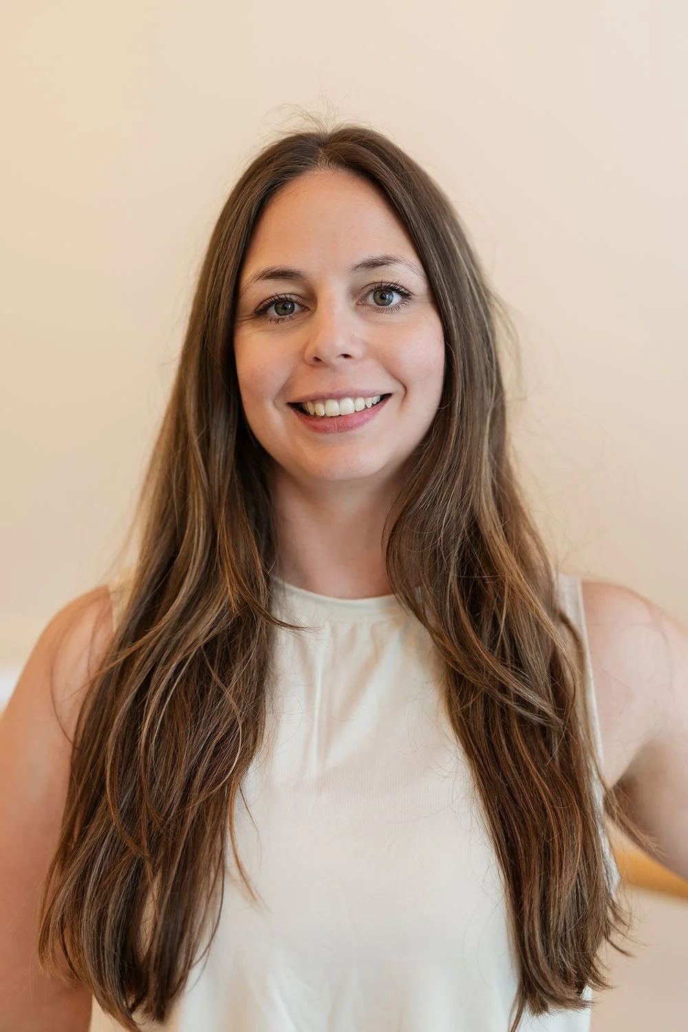 Portrait of a smiling woman with long brown hair wearing a sleeveless white top.