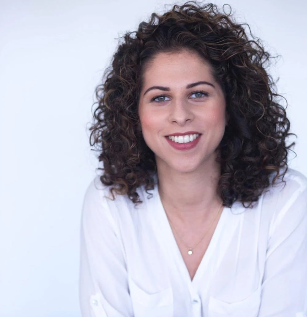 A woman with curly brown hair, blue eyes, and light skin smiling in front of a plain white background.