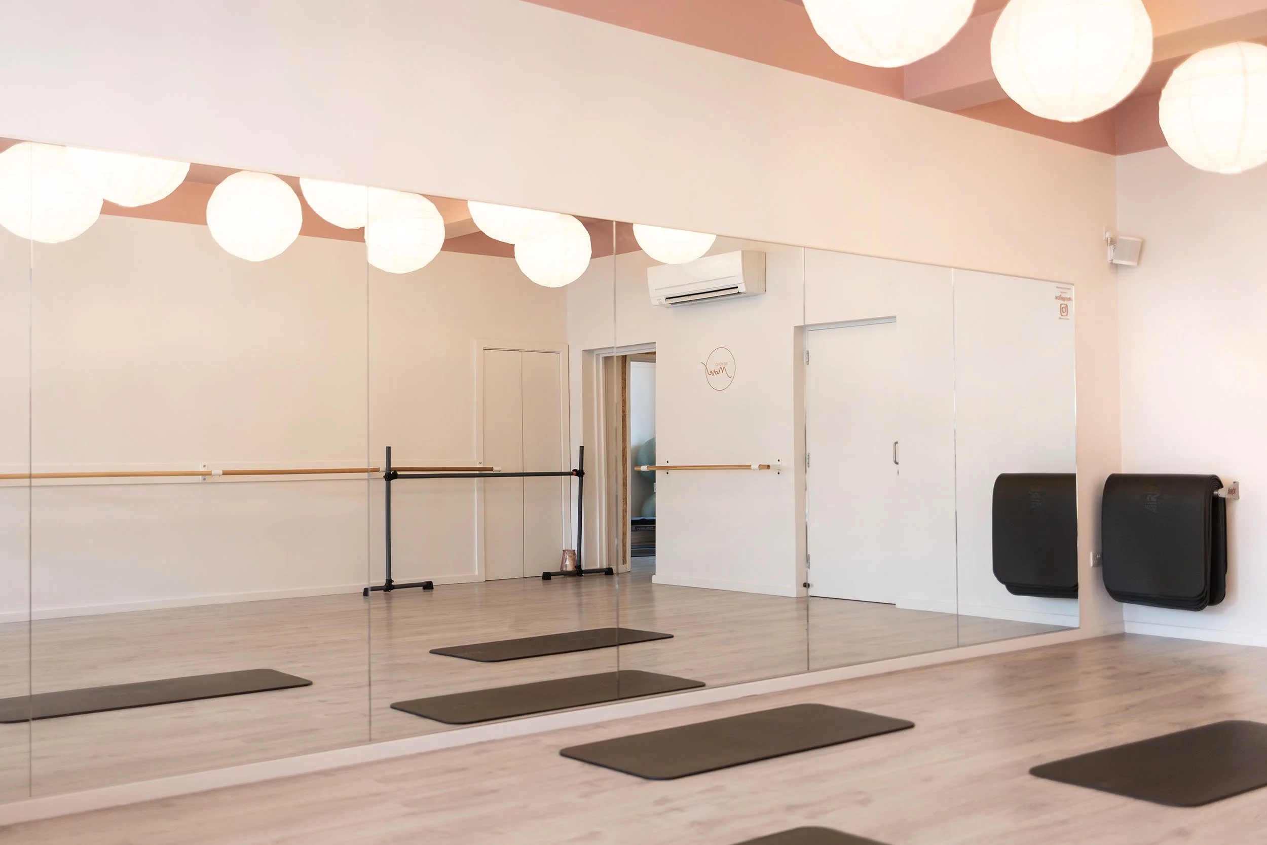 Empty dance or exercise studio with wall-mounted mirrors, yoga mats on the floor, ballet barre, wall-mounted black heater, and paper lantern ceiling lights.