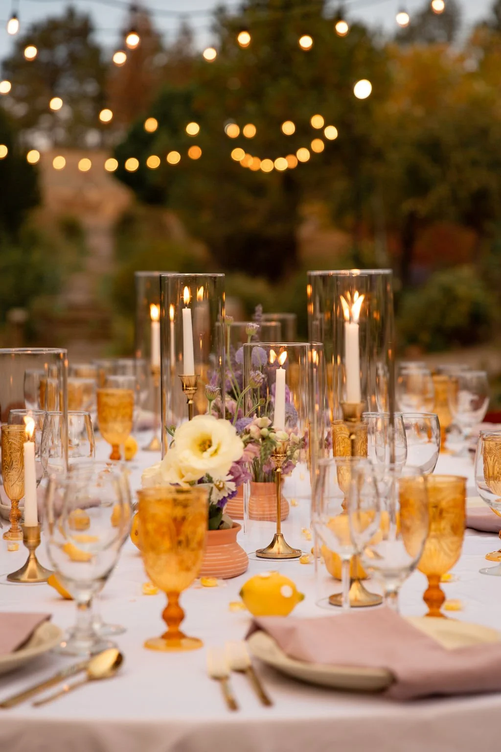 Elegant outdoor dinner table decorated with tall candleholders, glassware, flowers, and string lights in the background during evening.