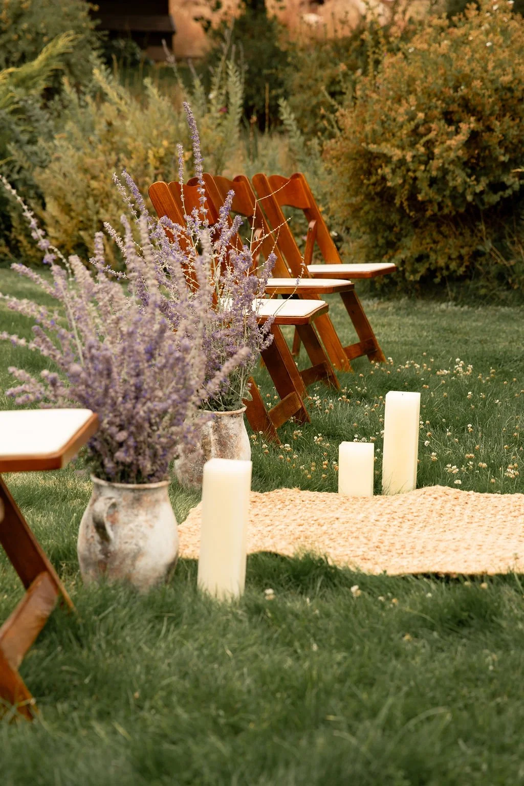 Outdoor wedding setup with wooden chairs, lavender flowers in pottery vases, white candles, and a woven rug on grassy ground, surrounded by lush bushes and trees.