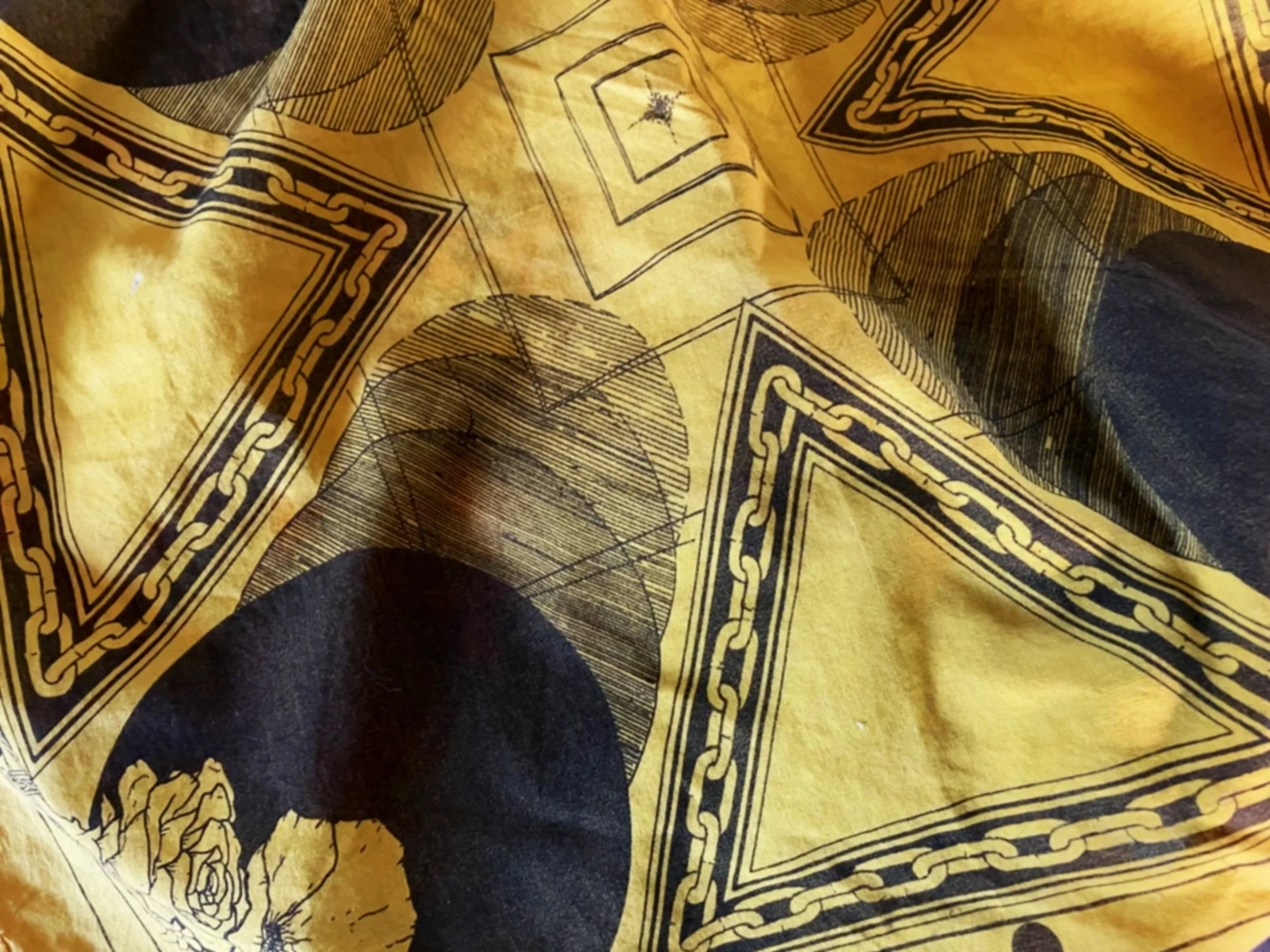Close-up of a yellow fabric with black printed design featuring chains, squares, a rose, and a large black circle.