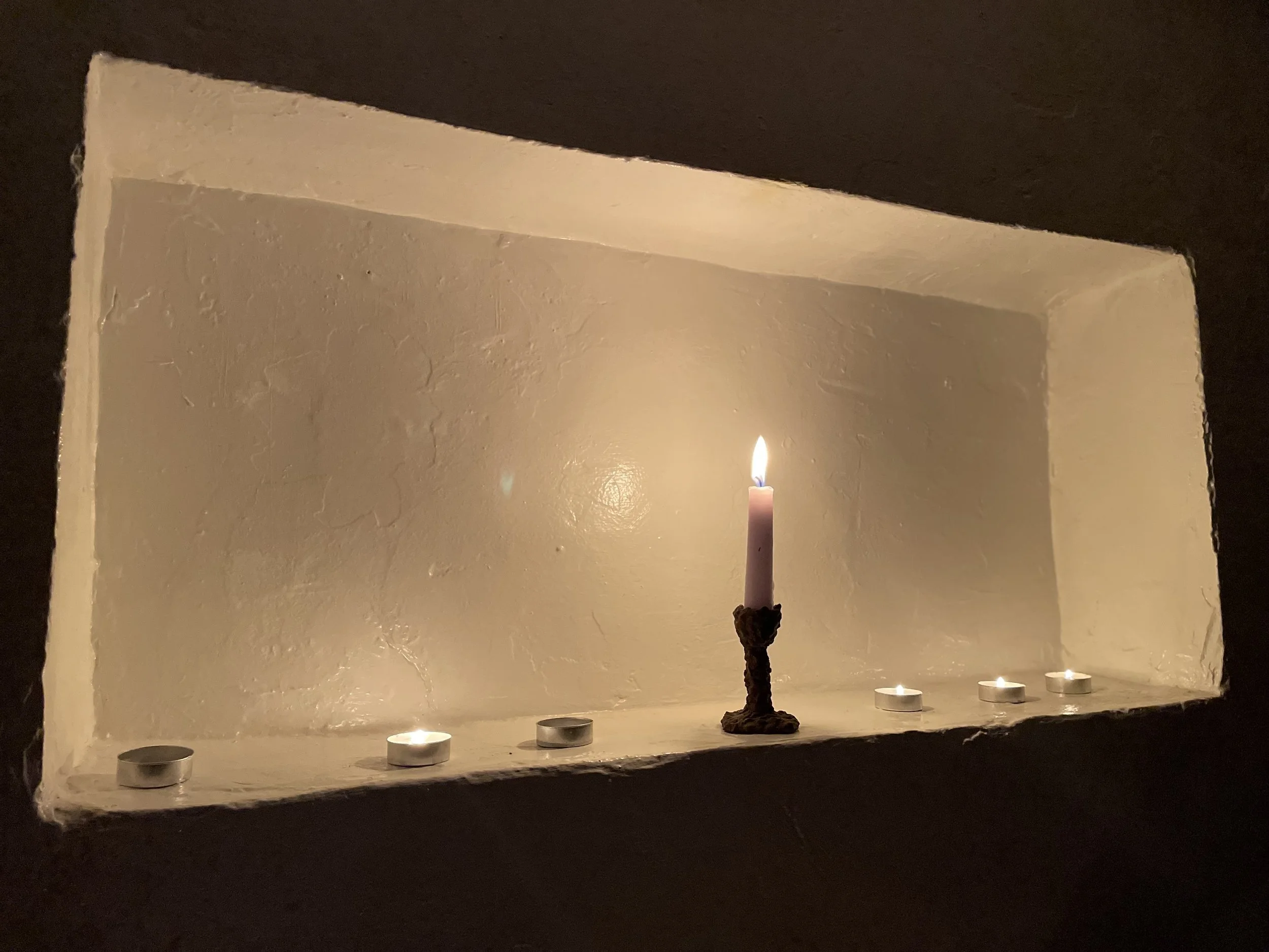 A small alcove with a white wall, containing one lit pink candle in a dark holder and four unlit tea light candles, with dark walls surrounding it.