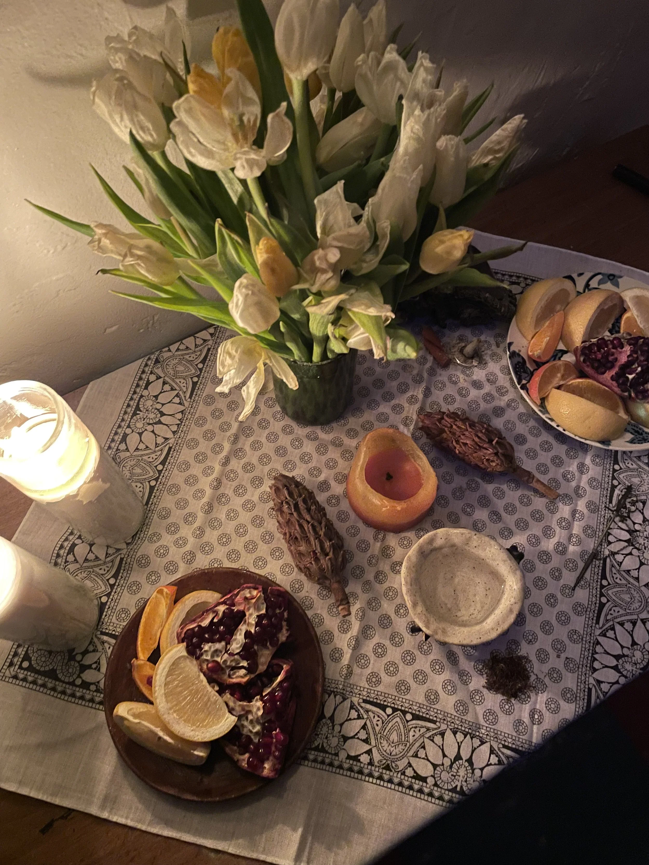 A table decorated with a bouquet of white and yellow tulips, a lit white candle, a plate of pomegranate, slices of lemon and orange, a horned owl figurine, a bowl, and a pomegranate in halves. The tablecloth has intricate black and white patterns.