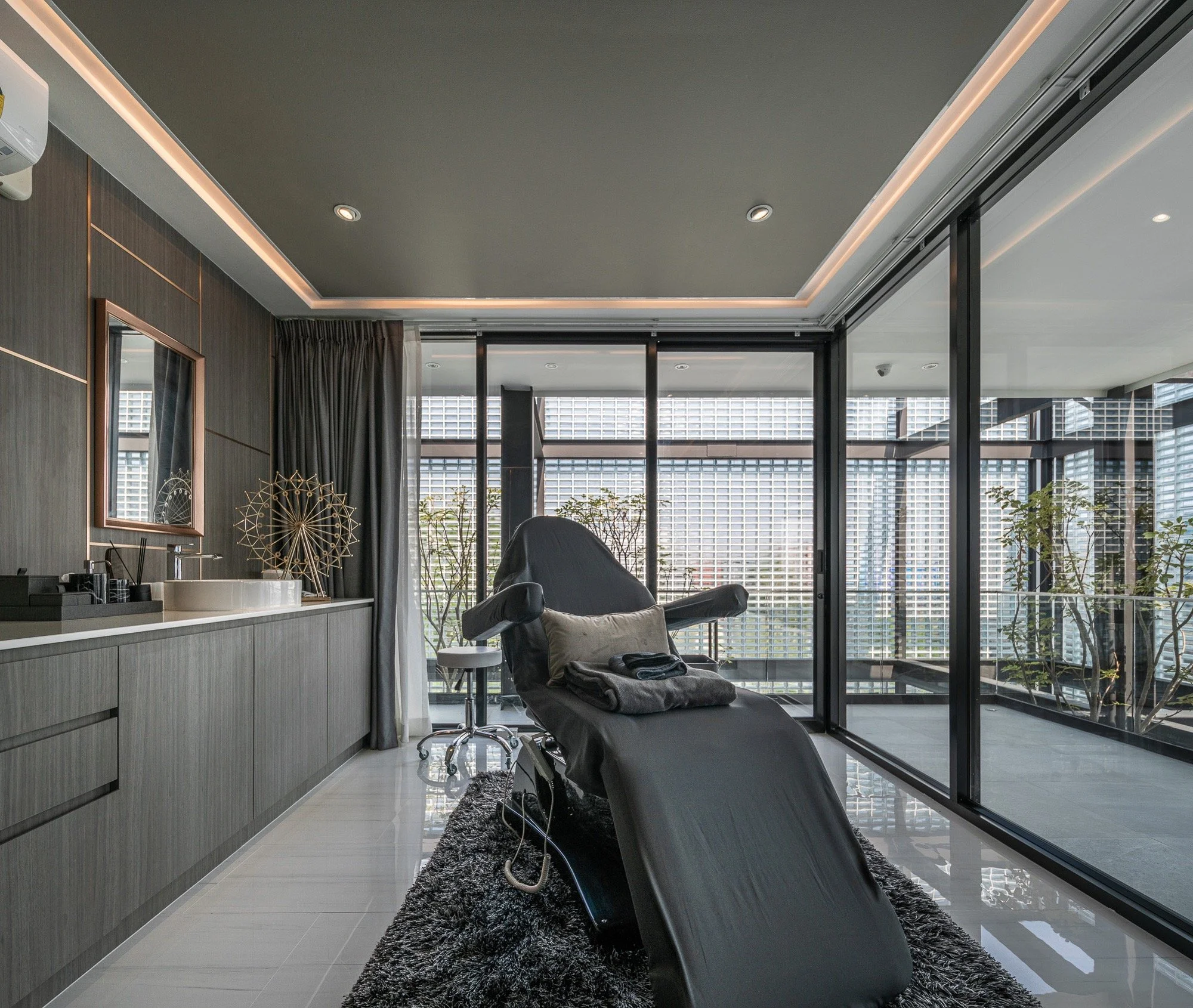 Modern medical or spa treatment room with black examination chair, large windows, and outdoor balcony with plants