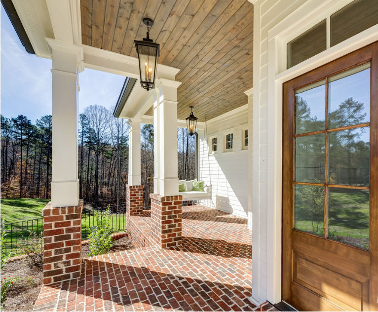 Covered porch of a home with brick flooring, white columns with brick bases, hanging lantern-style lights, a wooden door, and a white house with windows in the background, overlooking trees and a yard.