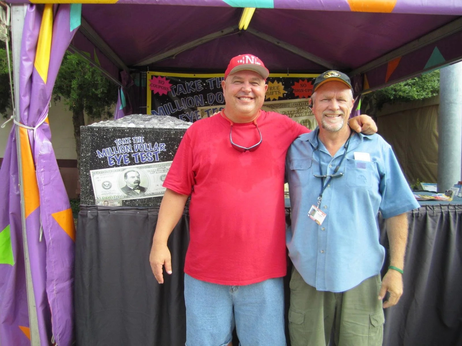 Two smiling men standing side by side in front of a booth with a sign that reads 'Take the Million Dollar Eye Test' and a large prop of a hundred dollar bill. The man on the left is wearing a red shirt and cap, and the man on the right is wearing a b