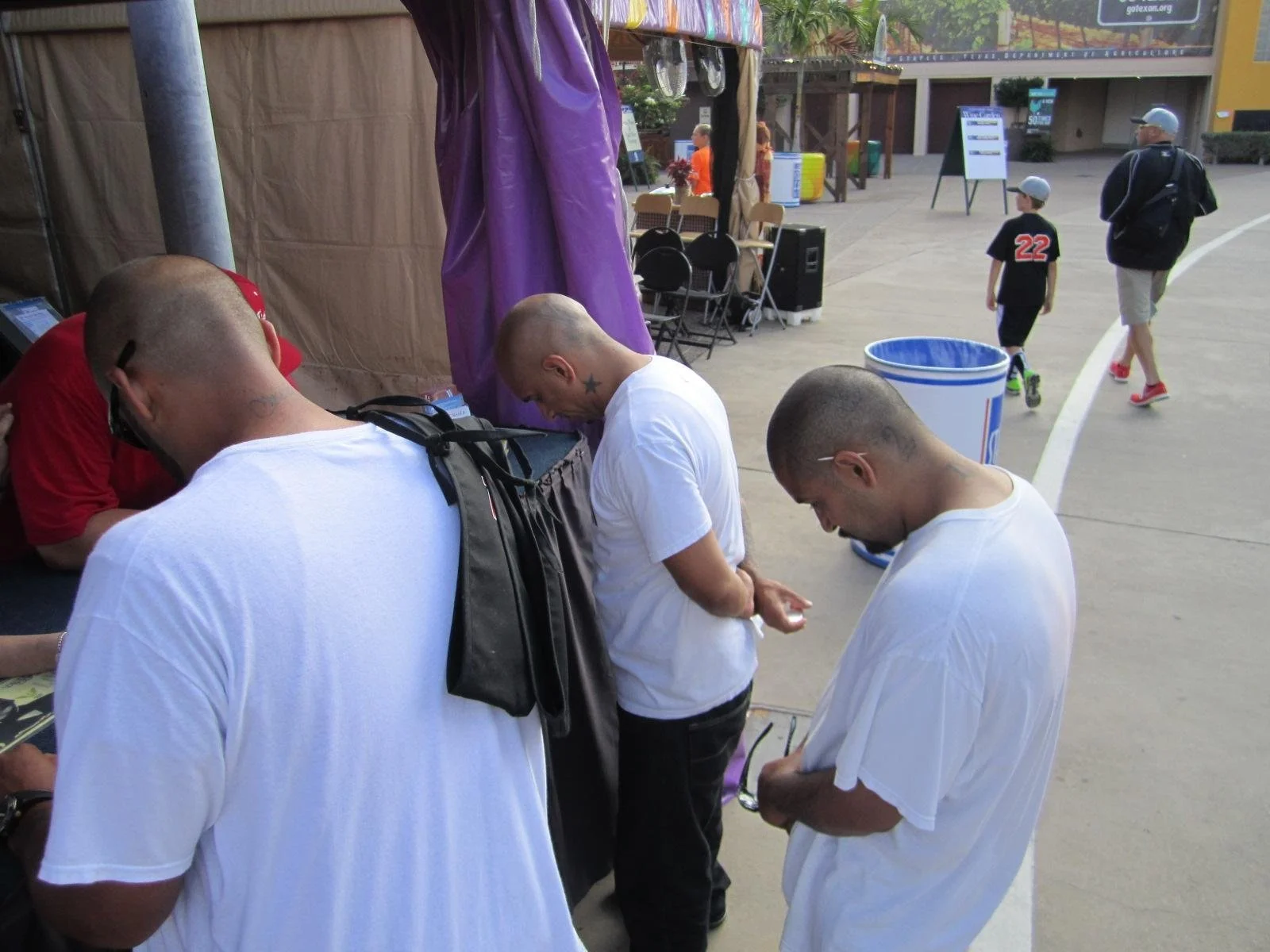 Three men with shaved heads and tattoos, wearing white T-shirts, are standing in line at an outdoor event, looking down at their phones or tickets. In the background, a man and boy are walking, dressed casually.