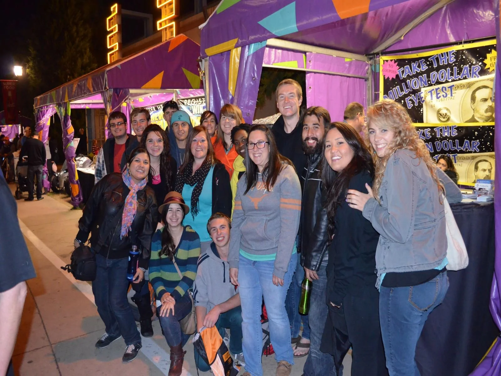 Group of smiling people standing together at a night outdoor event with purple tents and bright signs in the background.