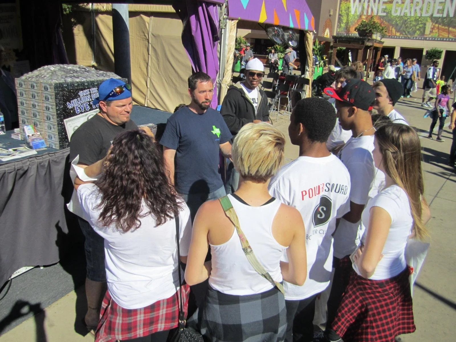 Group of people gathered outdoors at an event, some wearing white t-shirts, standing in front of a booth with a lottery display, with a colorful tent and a sign for a wine garden in the background