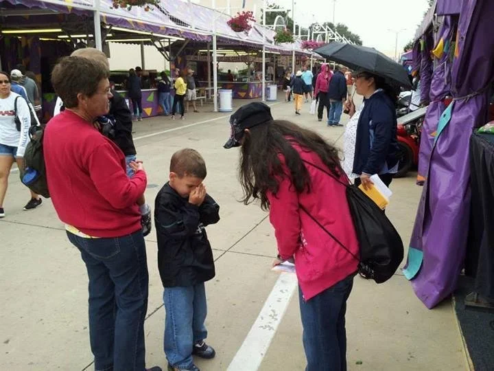 A woman with long hair, wearing a black cap and pink jacket, is bending down and talking to two children at an outdoor fair or carnival. The children appear emotional, with one wiping their face, while an older woman in a red sweater watches. In the