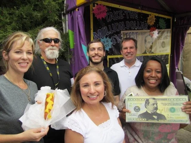 Group of six smiling people standing outdoors, holding a large fake hundred-dollar bill and a hot dog, with a colorful booth in the background.