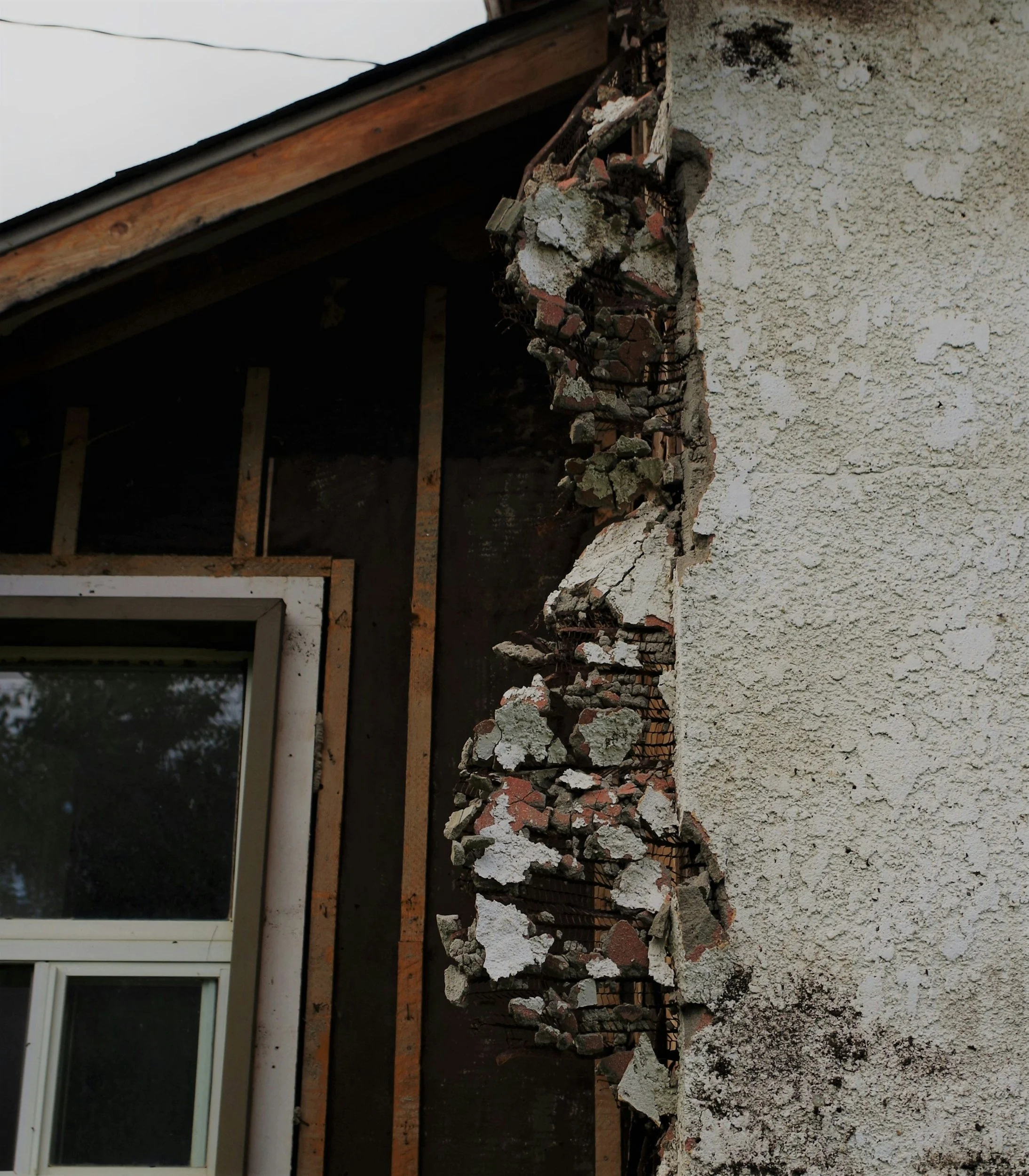 Close-up of a damaged wall with missing plaster exposing bricks, wires, and wooden framing behind the exterior surface.