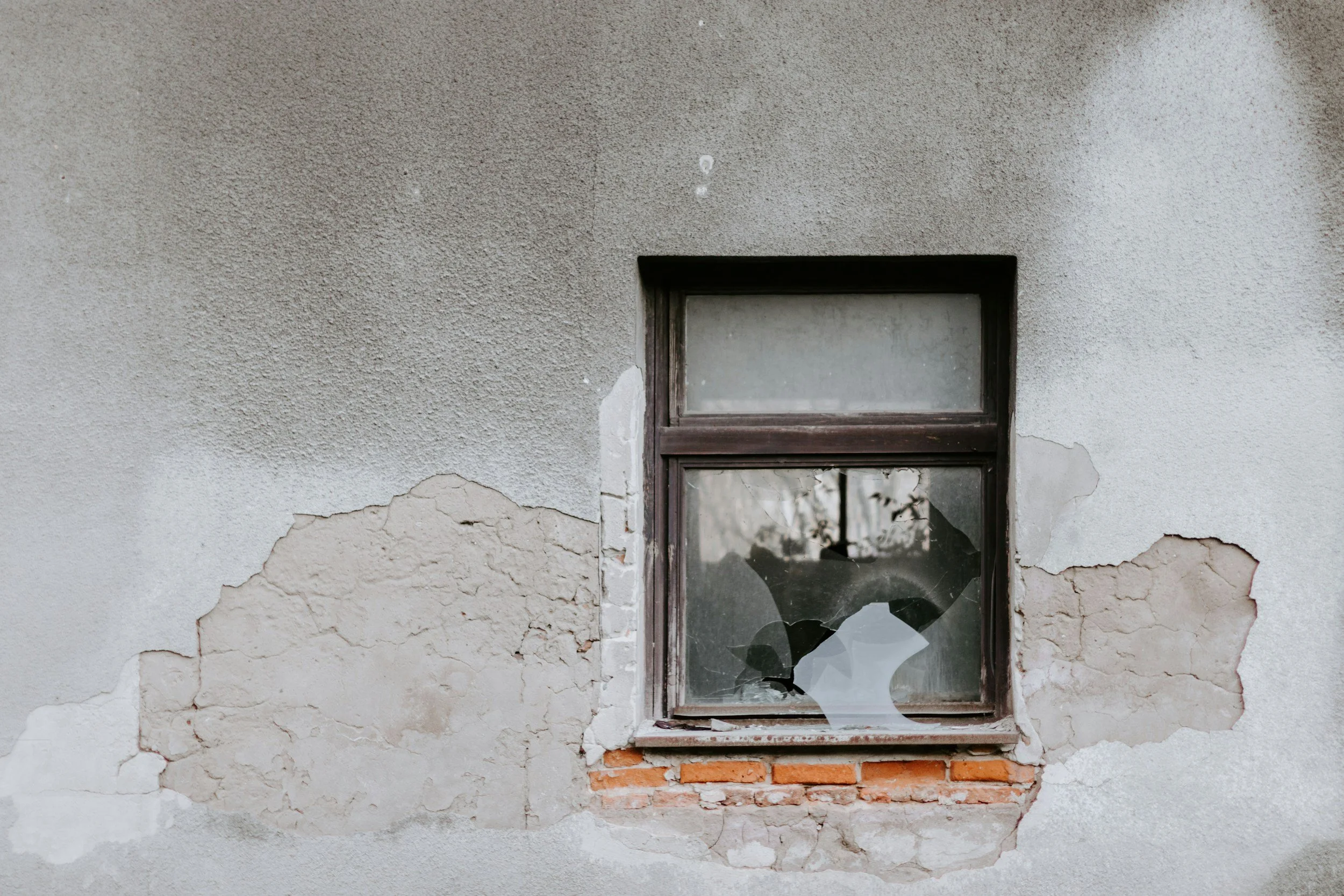 A cracked and partially peeling exterior wall with a broken window that has shattered glass, revealing reflections of trees and sky.