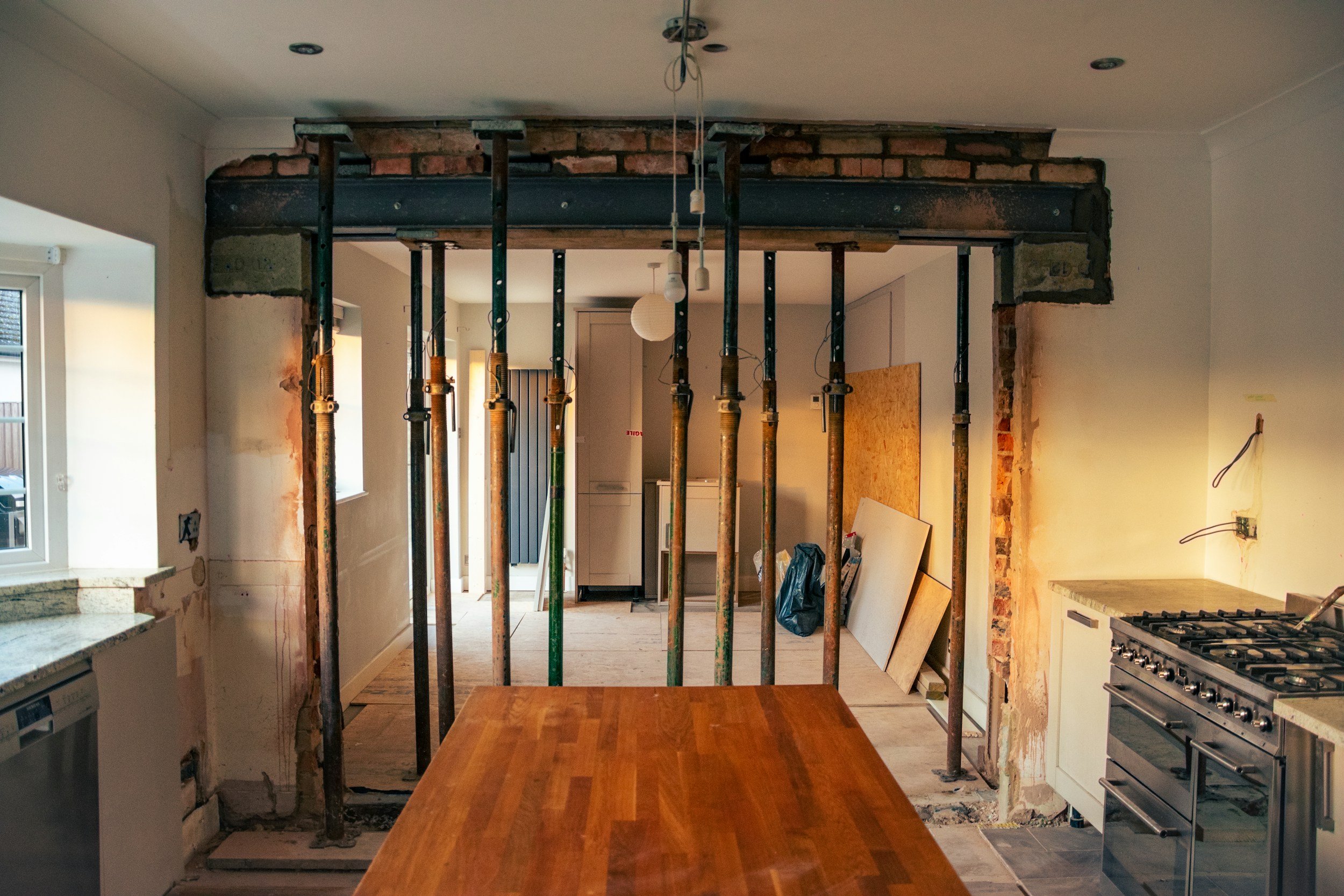 Kitchen under renovation with exposed brick and support poles, showing construction materials and partially removed walls.