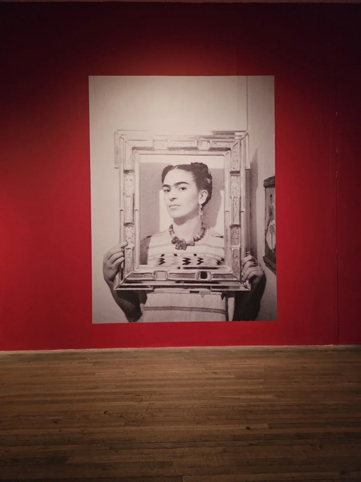 A black-and-white portrait of Frida Kahlo displayed against a red exhibition wall.