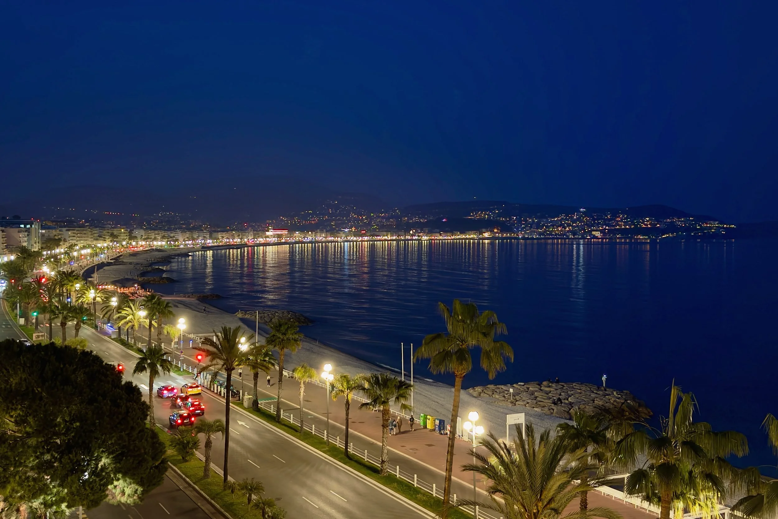Elevated night view of Nice’s curved bay with lit promenade palms reflecting in the Mediterranean