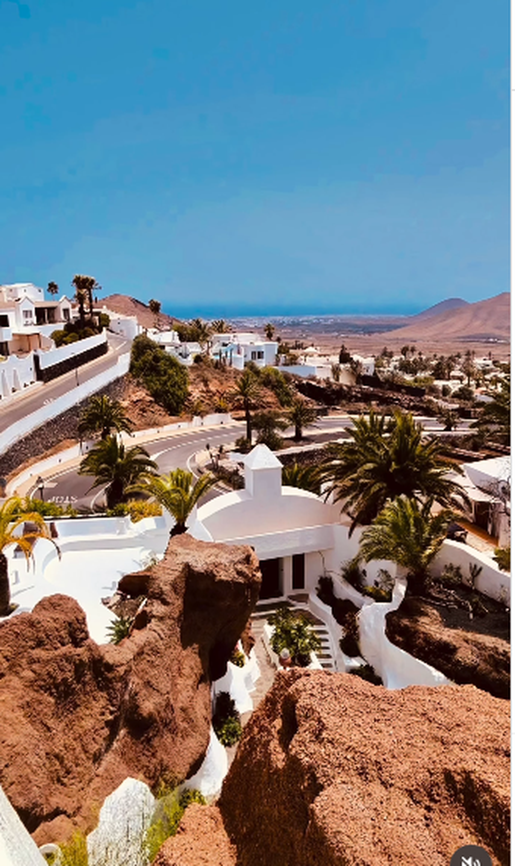 A scenic view of a hillside in a desert landscape with white buildings, palm trees, and a winding road, with mountains and the ocean in the distance under a clear blue sky.