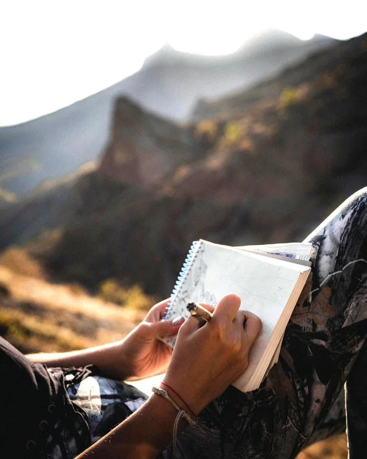 Une personne en vêtements de marche, assise dans un paysage montagneux, écrit dans un carnet avec un stylo, avec un paysage rocheux et montagneux en arrière-plan.