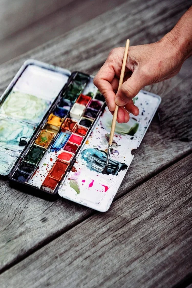 Une main tenant un pinceau plongeant dans une palette de peinture à l'aquarelle sur une table en bois.