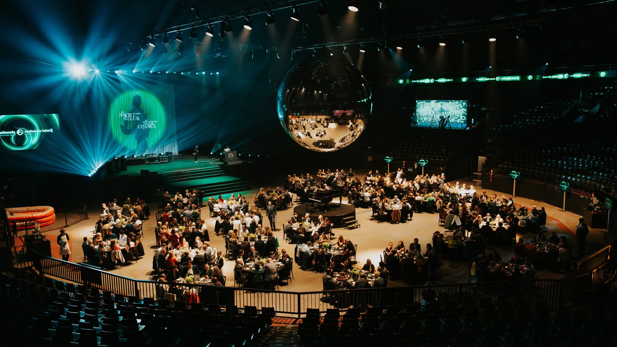 A large indoor event space with tables where people are dining. On the stage, there is a large reflective disco ball hanging from the ceiling, with spotlights and stage lighting creating a vibrant atmosphere. Multiple screens and banners display grap