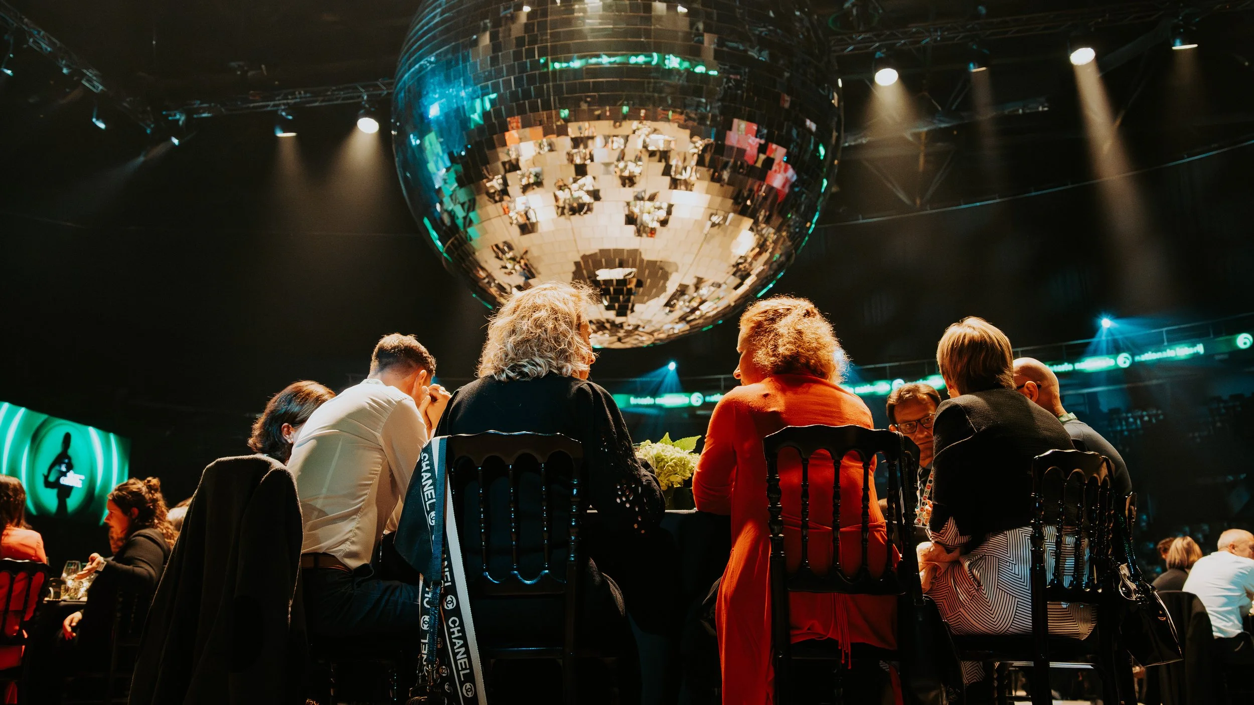 Group of people seated at tables in front of a large disco ball in a dark venue, with stage lights and screens in the background.