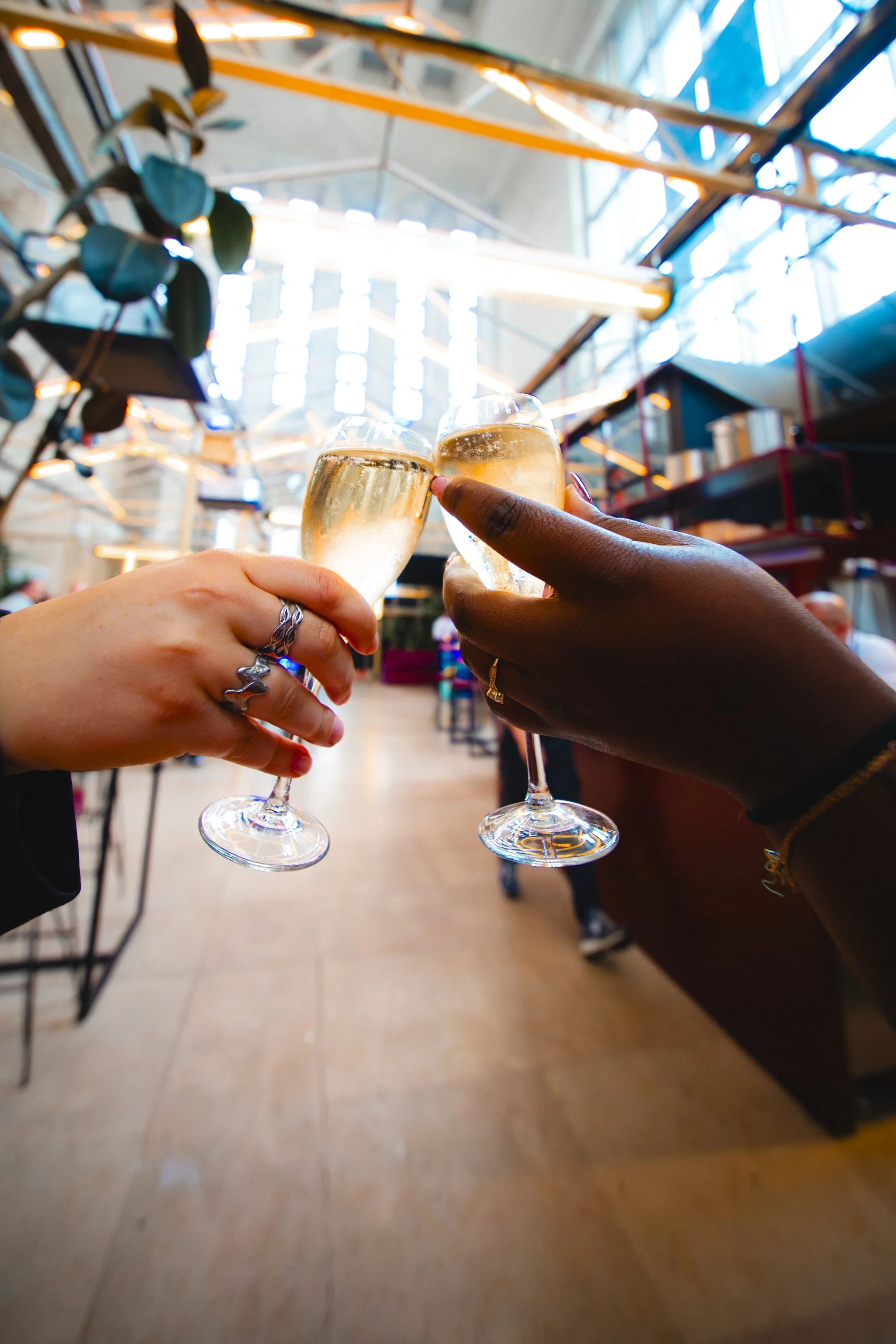 Two people holding champagne glasses in a toast inside a modern, brightly lit building with large glass windows and a high ceiling.
