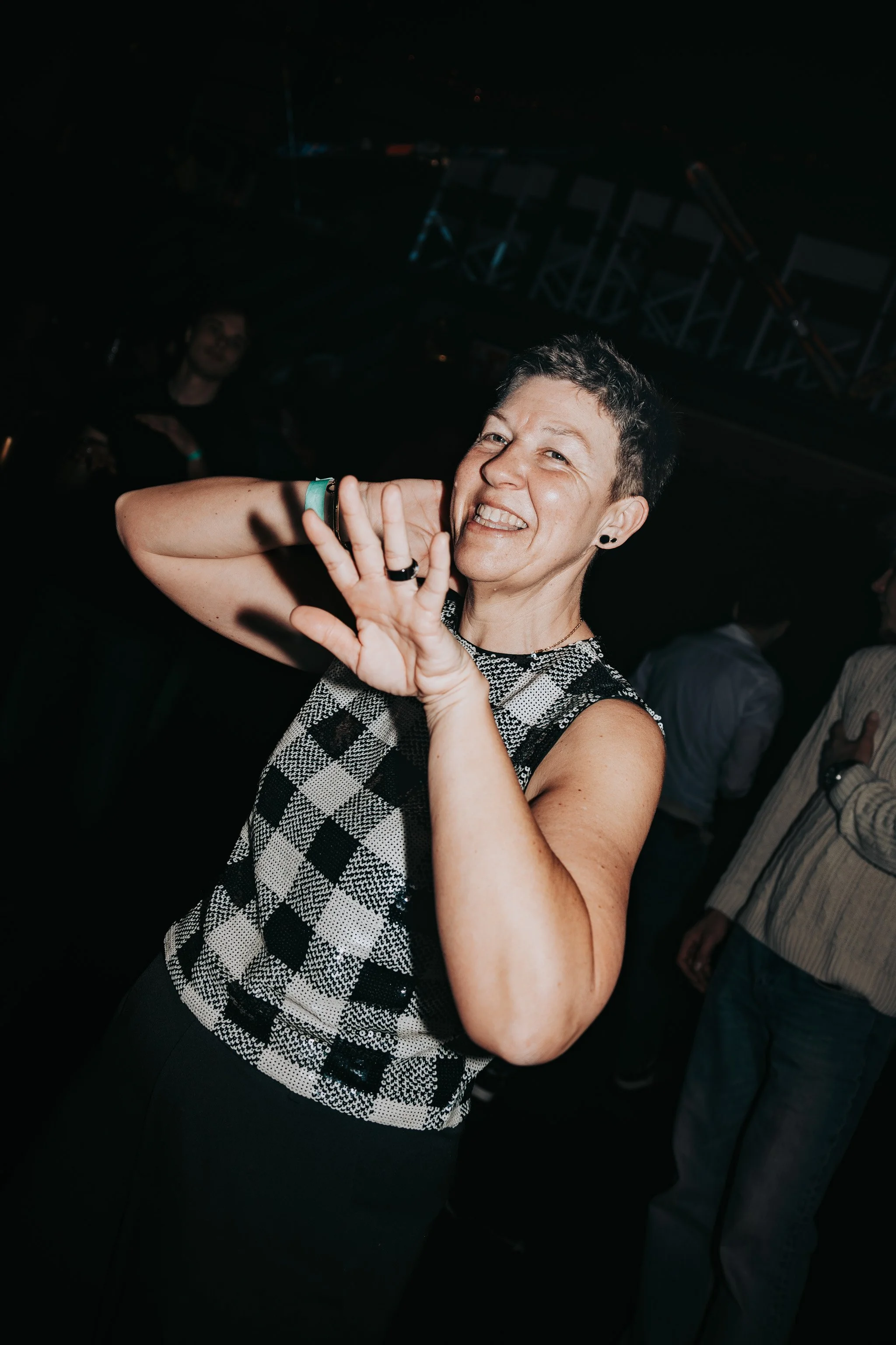 A woman smiling and making a peace sign with her right hand at a social event or party, with dark lighting and some people visible in the background.