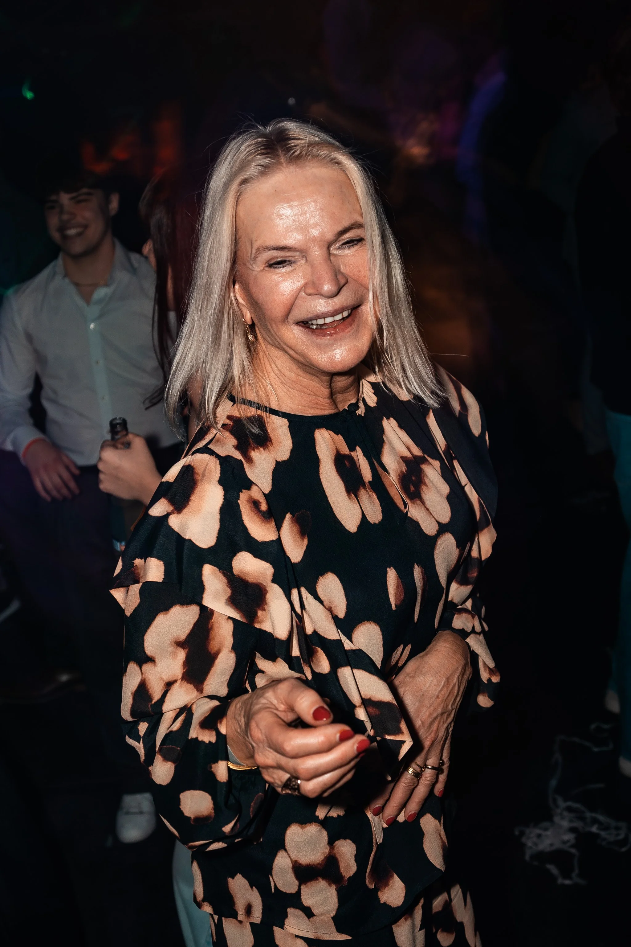 A smiling elderly woman with shoulder-length gray hair, wearing a leopard print blouse, holding a drink in her right hand, in a crowded dance or party scene with people in the background.