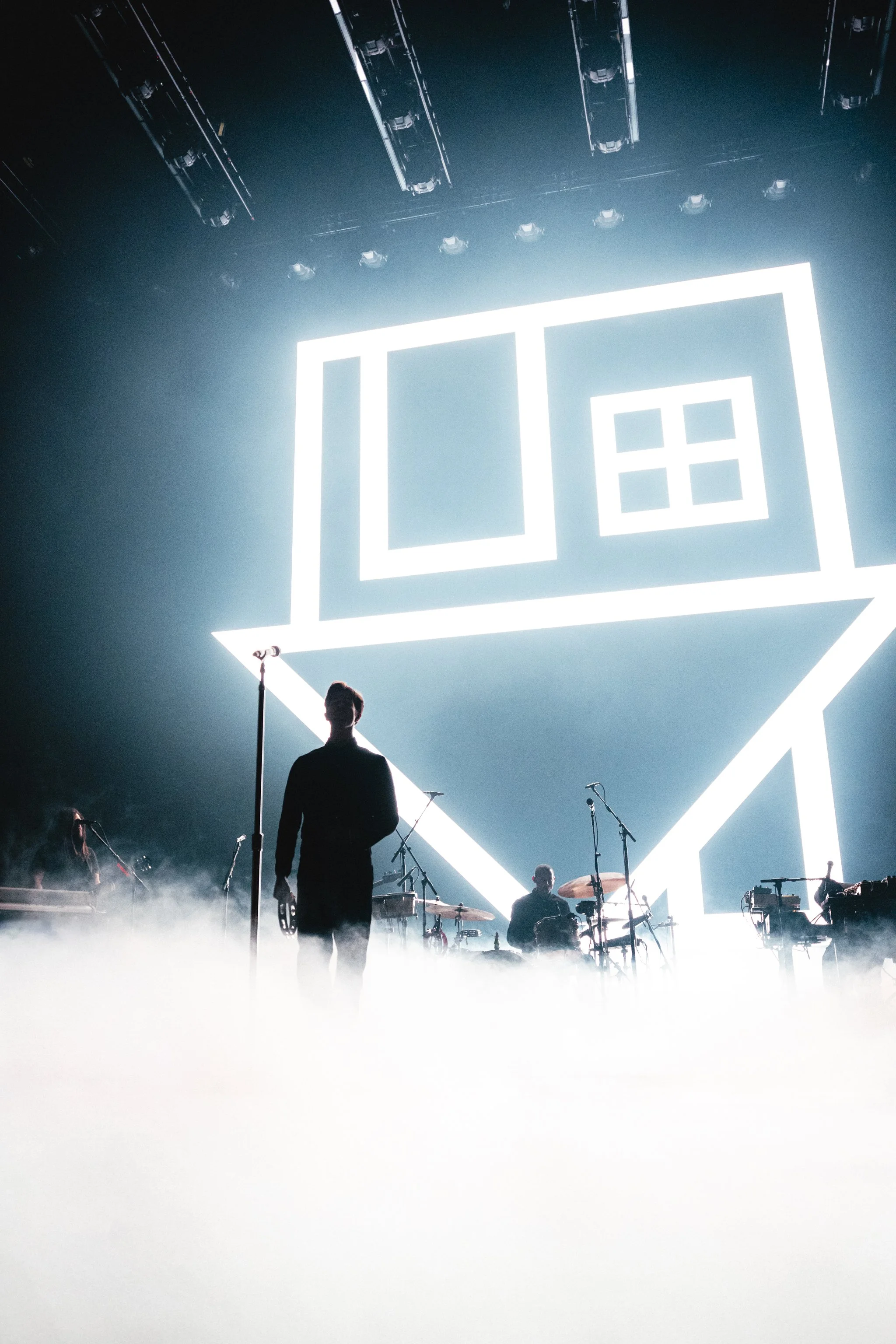 Silhouette of a person standing on stage with musical instruments, in front of a large, illuminated geometric logo, surrounded by stage lights and fog.