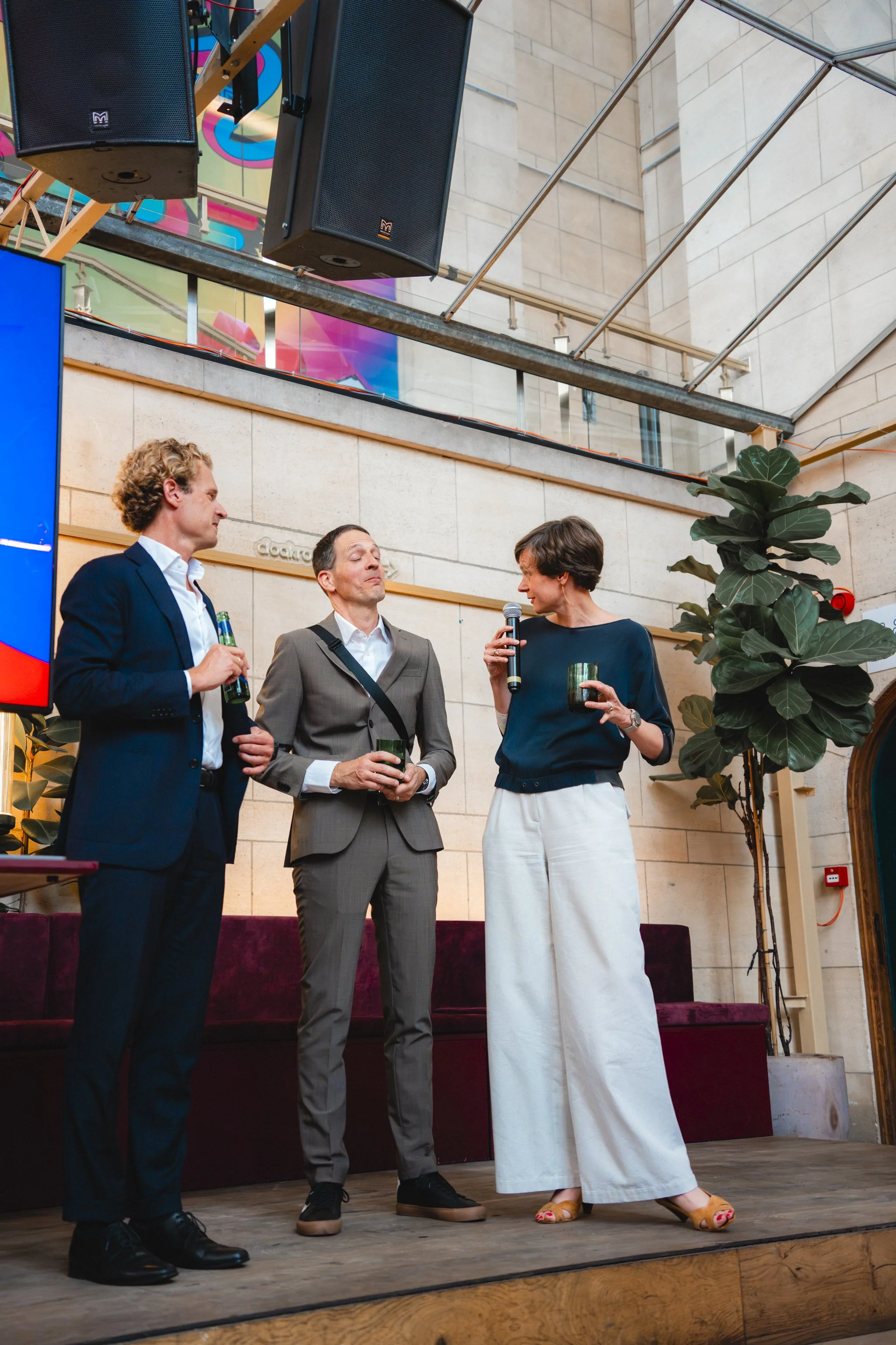 Three people standing on a stage, engaged in conversation. A man in a navy suit and white shirt holds a drink. A man in a gray suit with a crossbody bag holds a drink. A woman with short hair, in a navy top and white pants, holds a microphone and a d