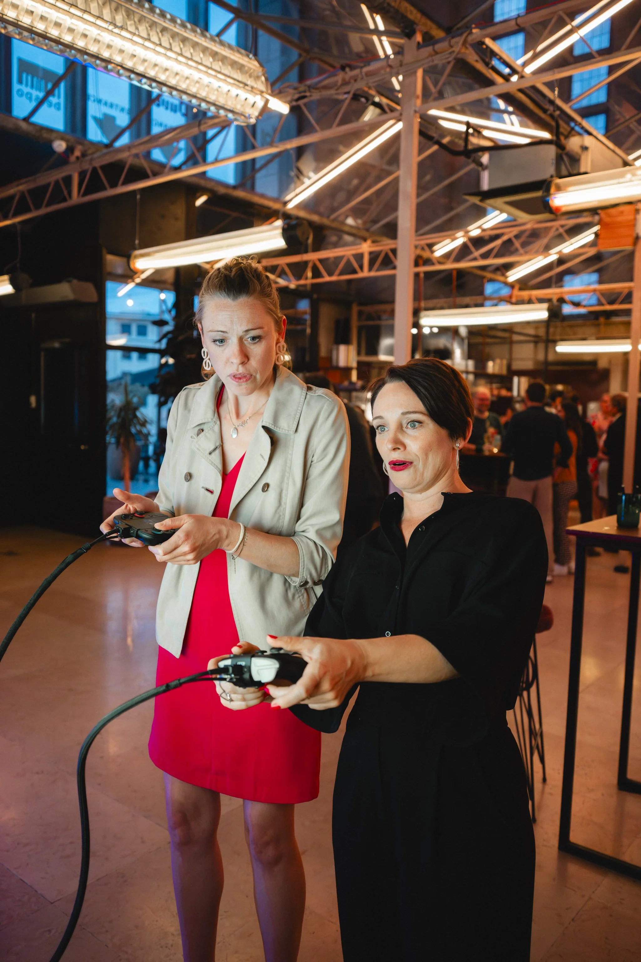 Two women are standing in a modern indoor space with exposed ceiling beams and industrial lighting. They are looking at and holding gaming controllers, possibly playing a video game. The woman on the left wears a red dress and a beige blazer, and the