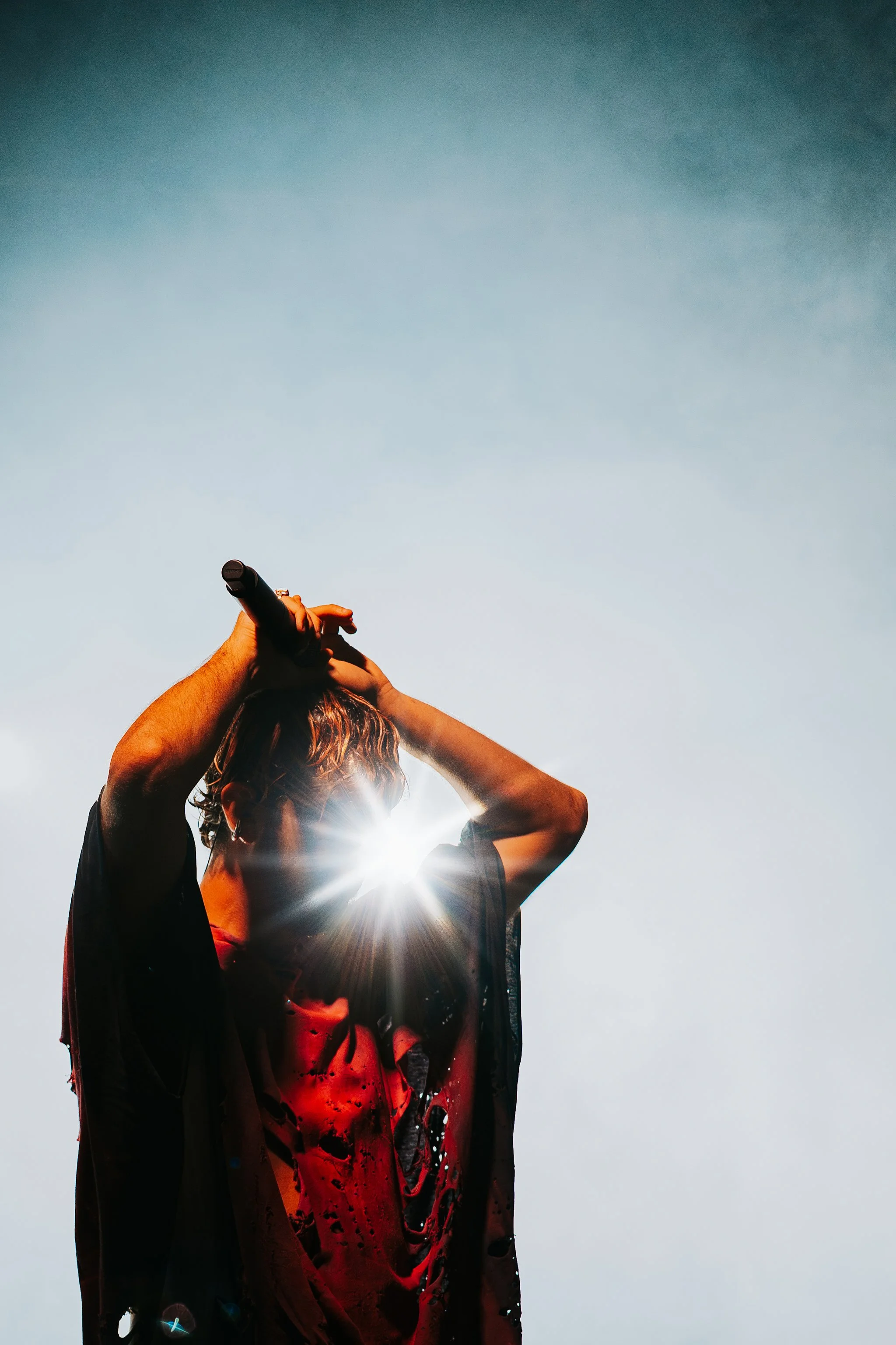 Person with curly hair holding a microphone up to their mouth, backlit by the sun, standing outdoors against a clear sky.