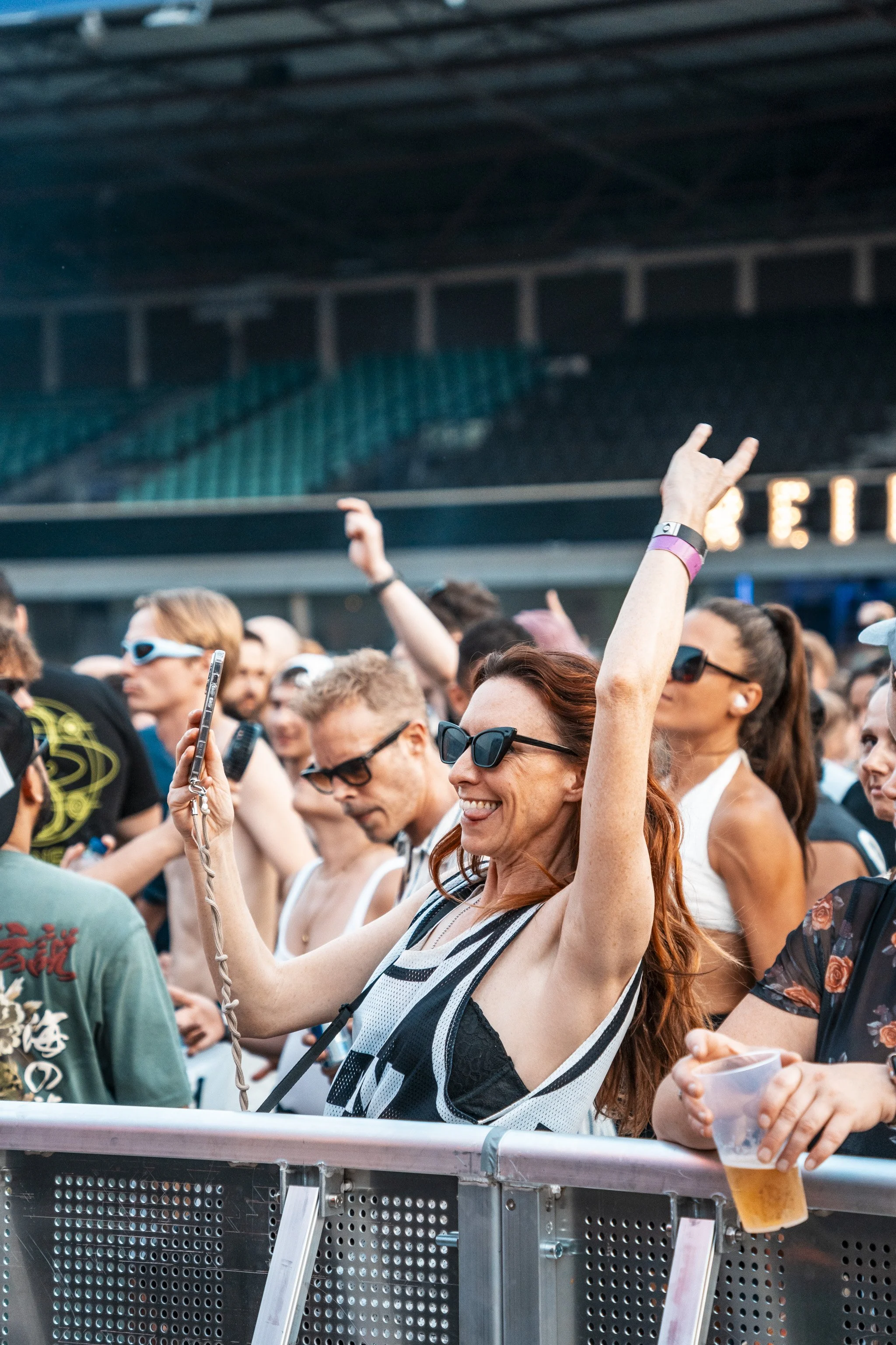 A group of people at an outdoor music event, with one woman in the foreground making a peace sign and holding a plastic cup of beer, all wearing casual summer clothing and sunglasses.