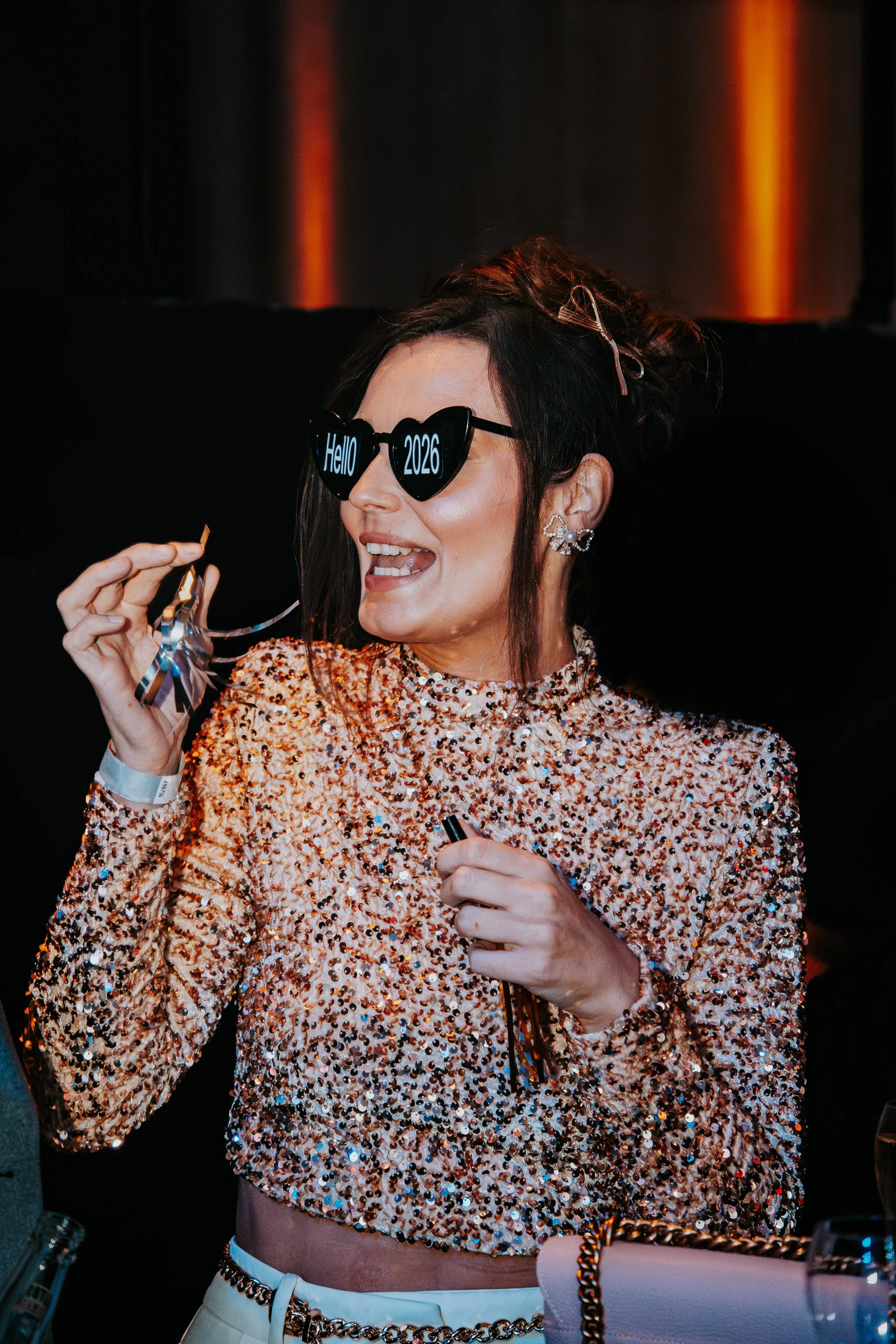 A woman wearing heart-shaped sunglasses that say 'Hello 2026,' holding a party blower, dressed in a sequined top, with earrings, a bracelet, and surrounded by festive decorations in a dark setting.