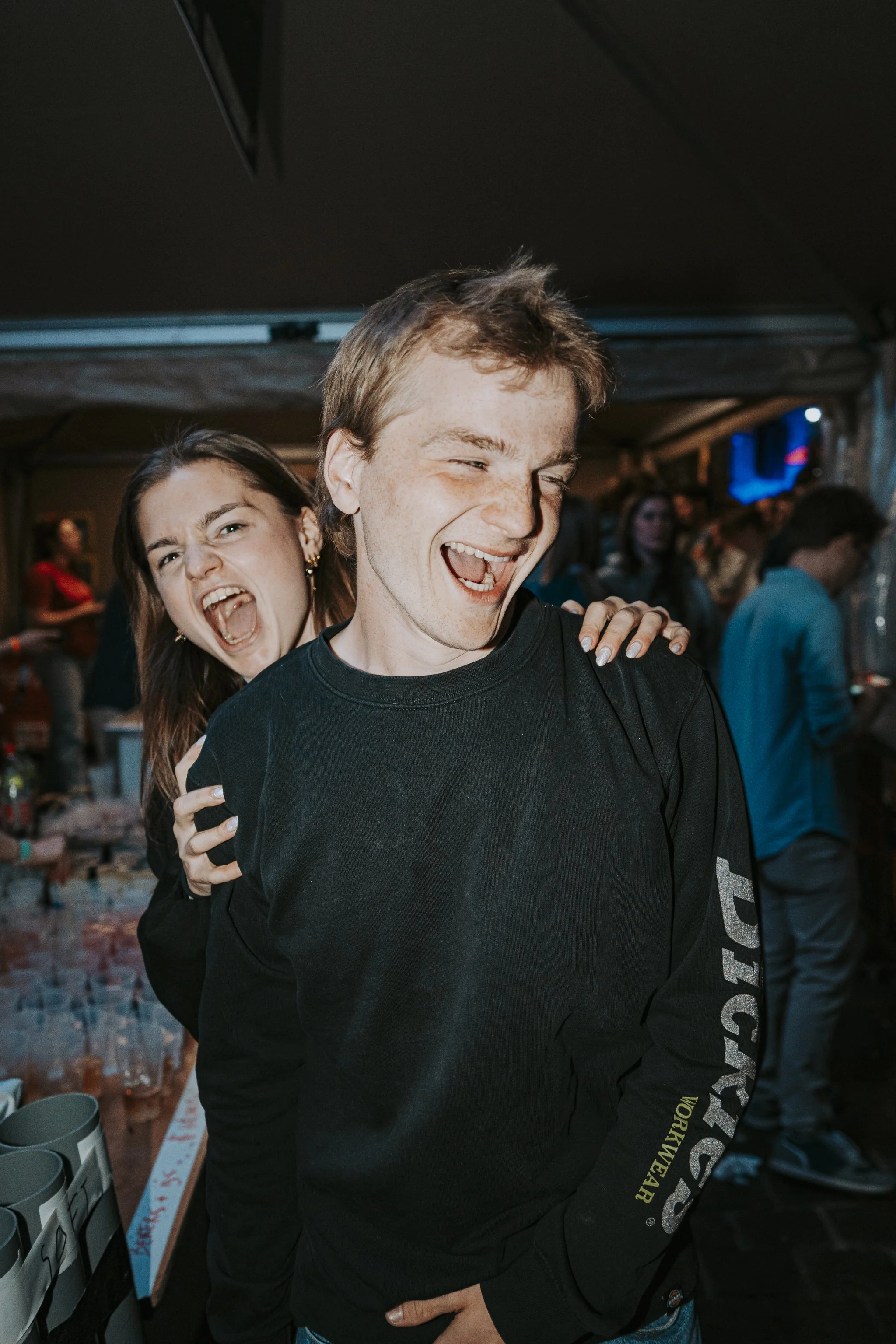 Two young adults, a woman and a man, are smiling and having fun at a lively social gathering or party; the woman is behind the man with her arm on his shoulder, and both are happy and laughing.