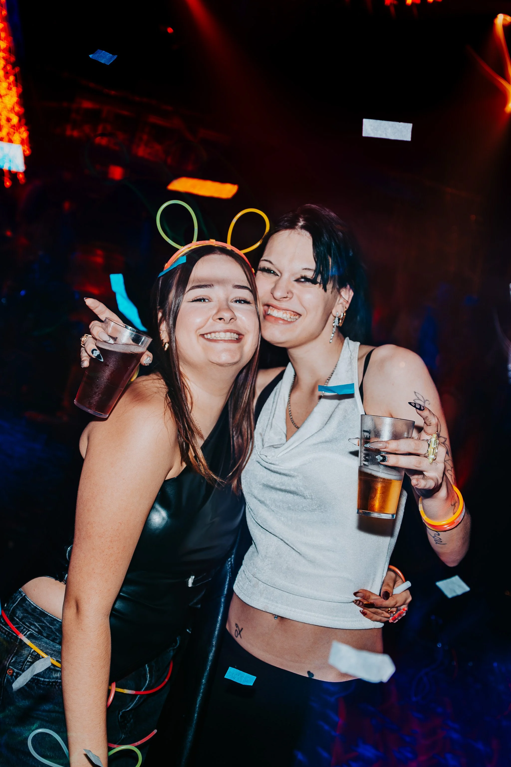 Two women smiling and hugging each other at a party, holding drinks and wearing colorful glow jewelry and bunny ears headbands with confetti in the background.
