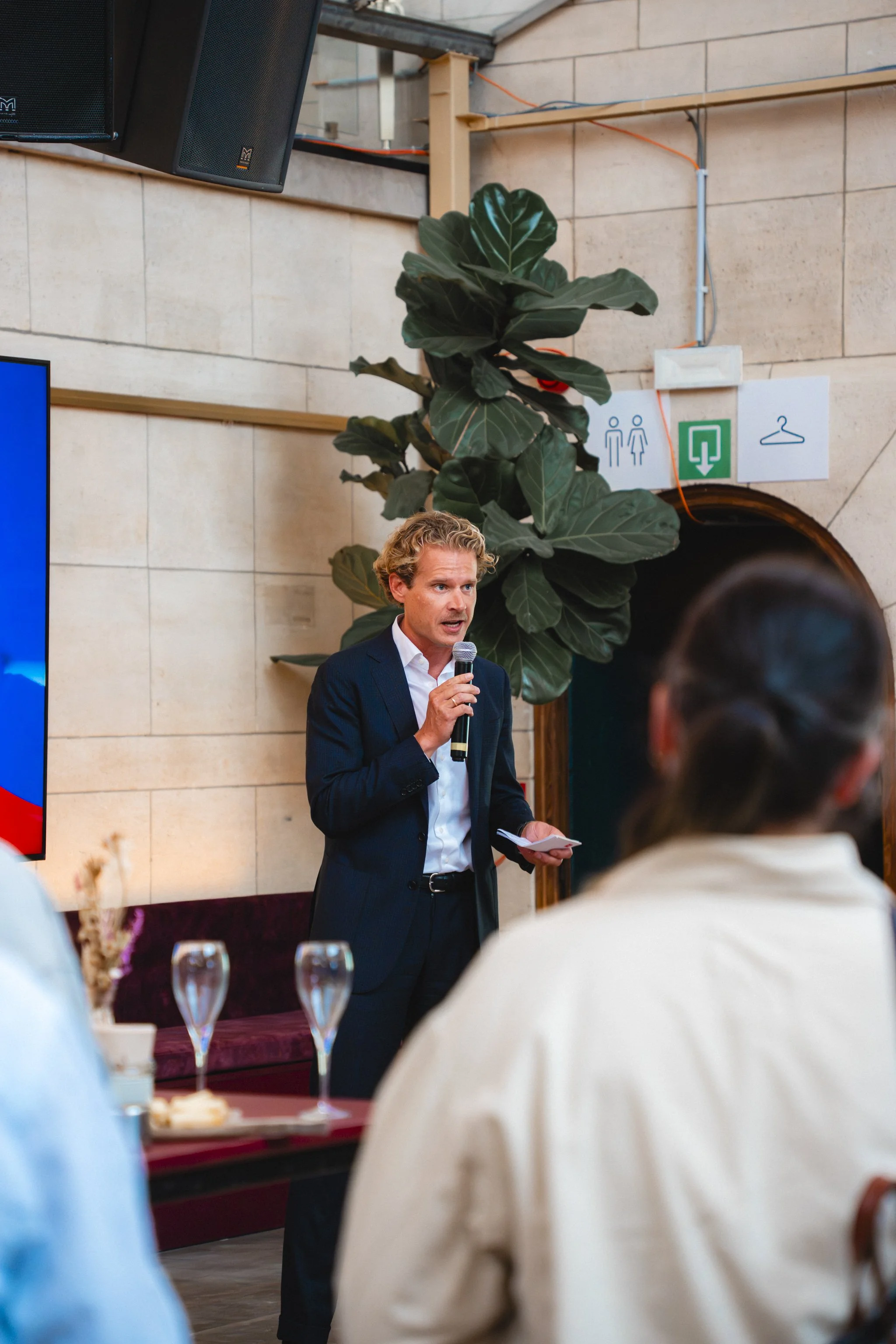 A man in a suit holding a microphone and notes, speaking at a formal event in a room with beige stone walls, large plants, and emergency signs.