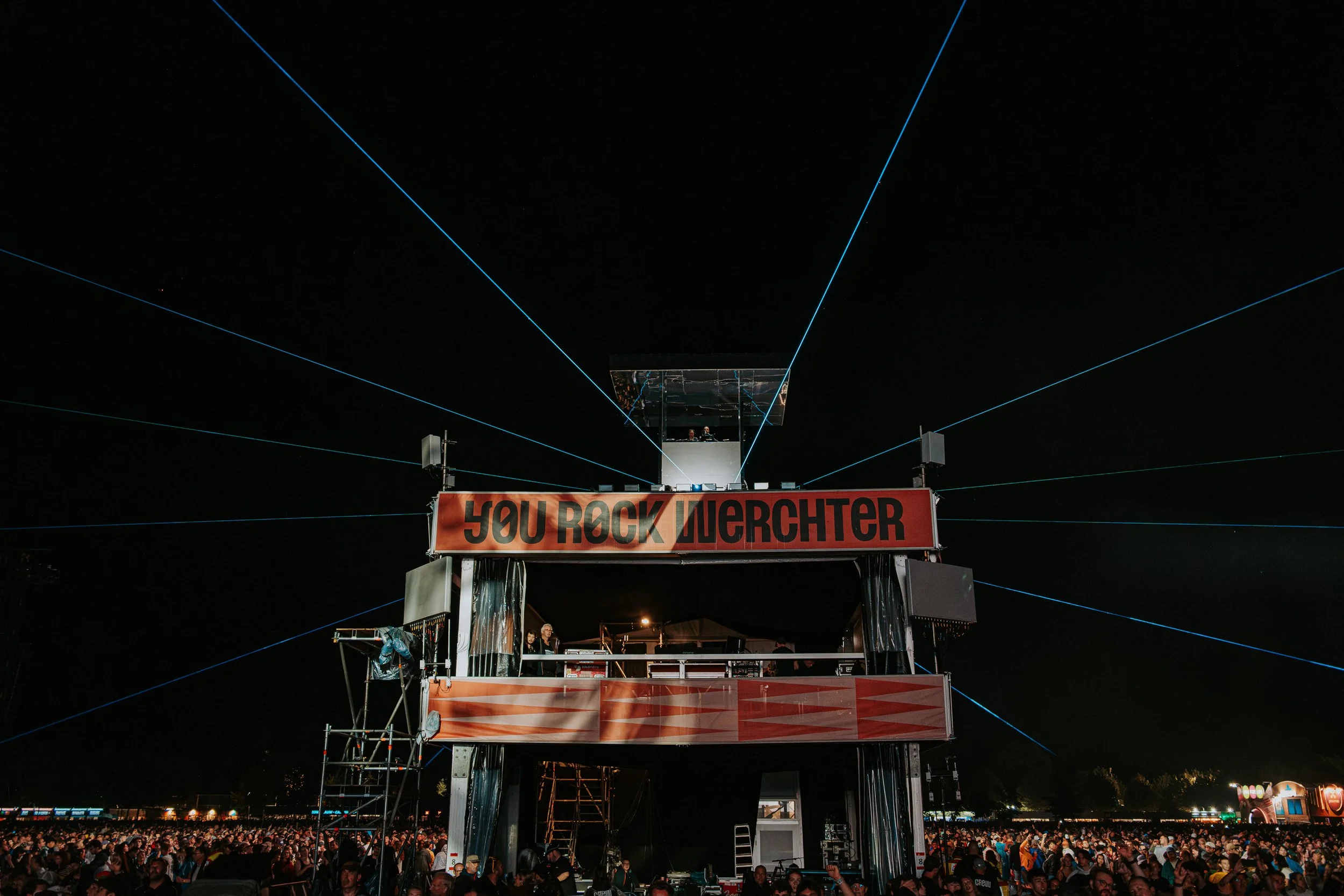 Nighttime scene at a music festival with a large crowd. A stage with a sign that reads "YOU ROCK WUERCHTER" is illuminated, with blue laser lights projecting outward into the dark sky above the crowd.