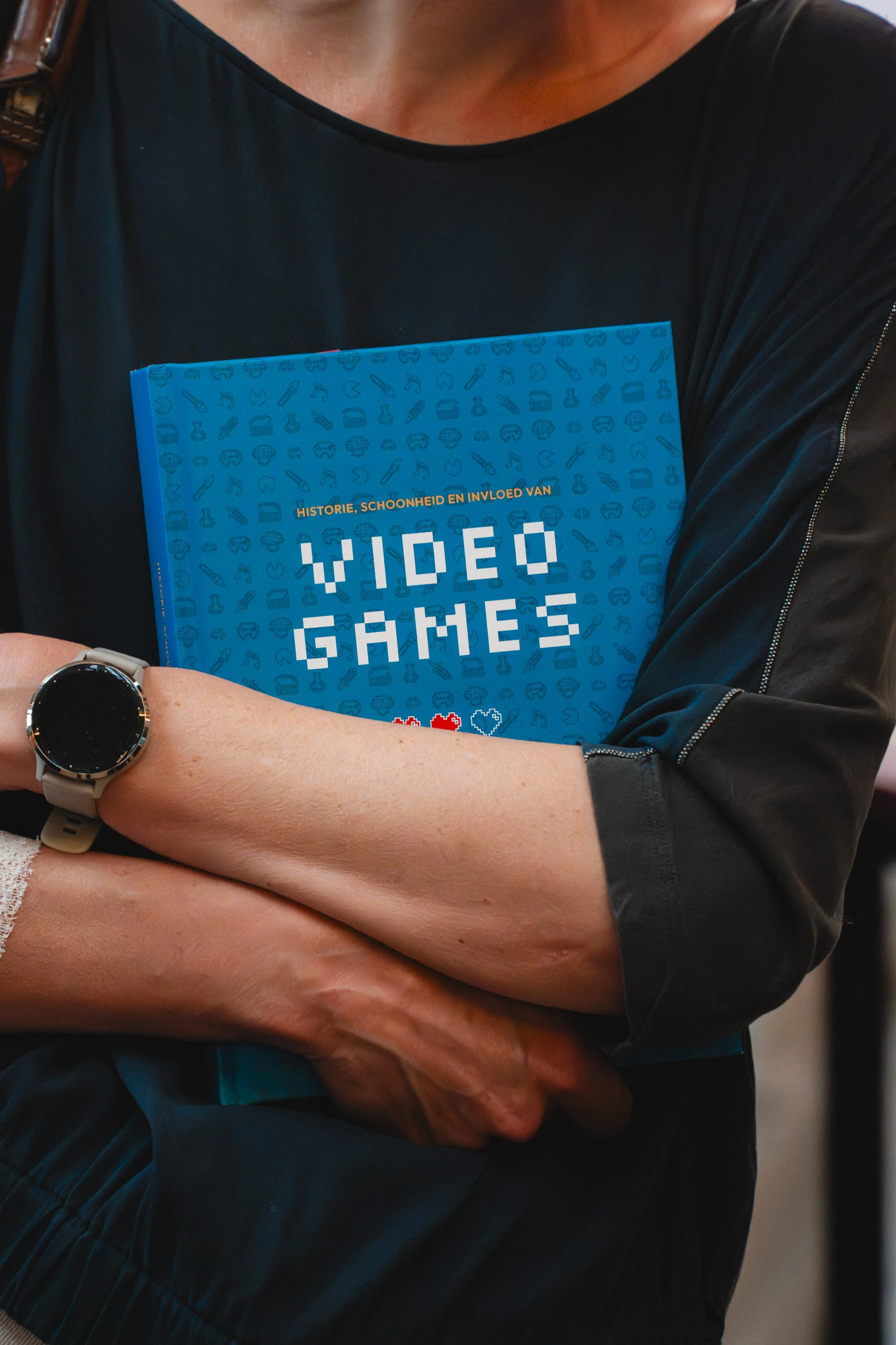 Person standing with arms crossed, holding a copy of a blue book titled 'Video Games' with icons of various gadgets and symbols related to technology and gaming.