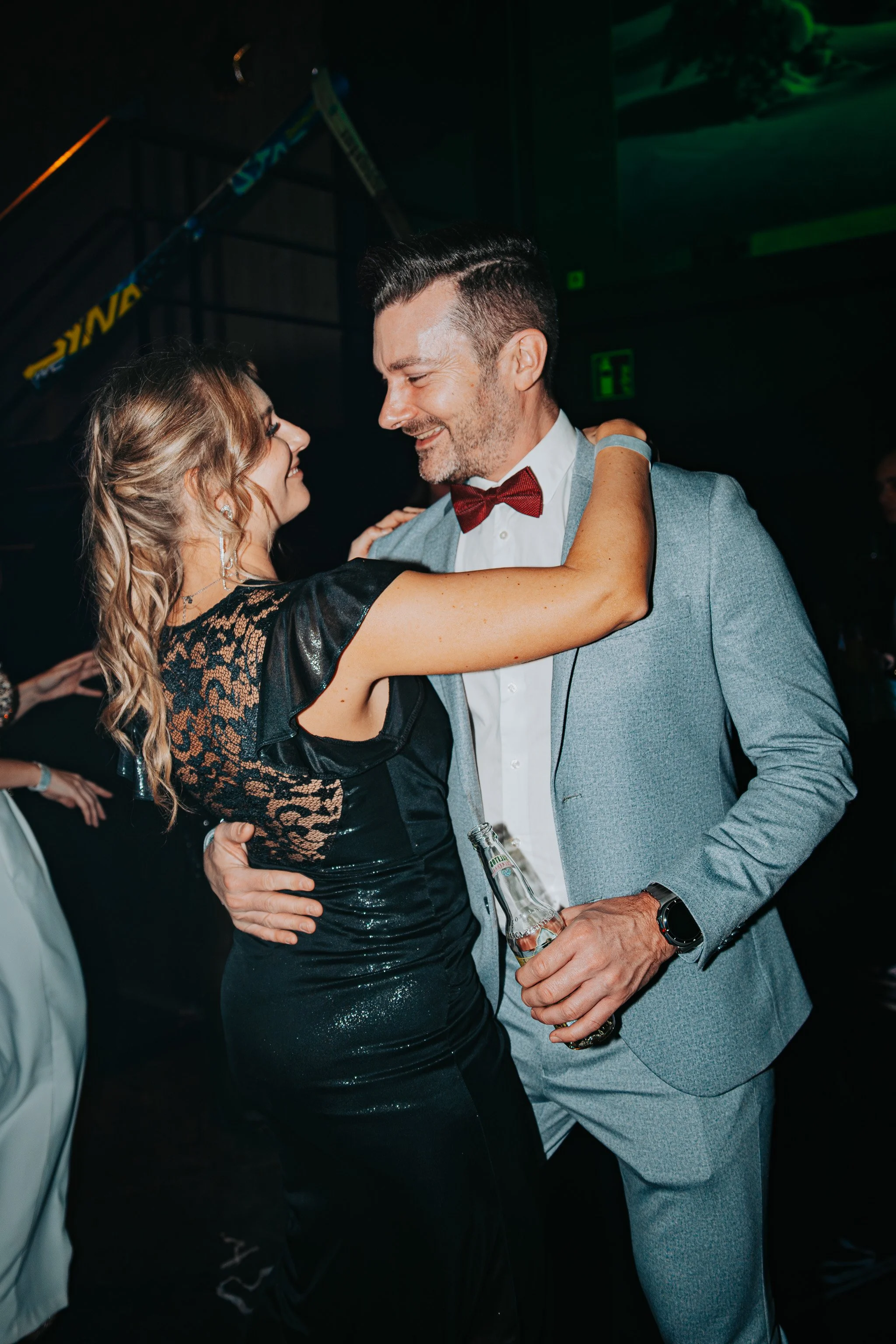 A man and woman dancing closely at a party, both smiling, with the woman wearing a black, lacy dress with sparkles and the man in a light gray suit with a red bow tie, holding a beer bottle.