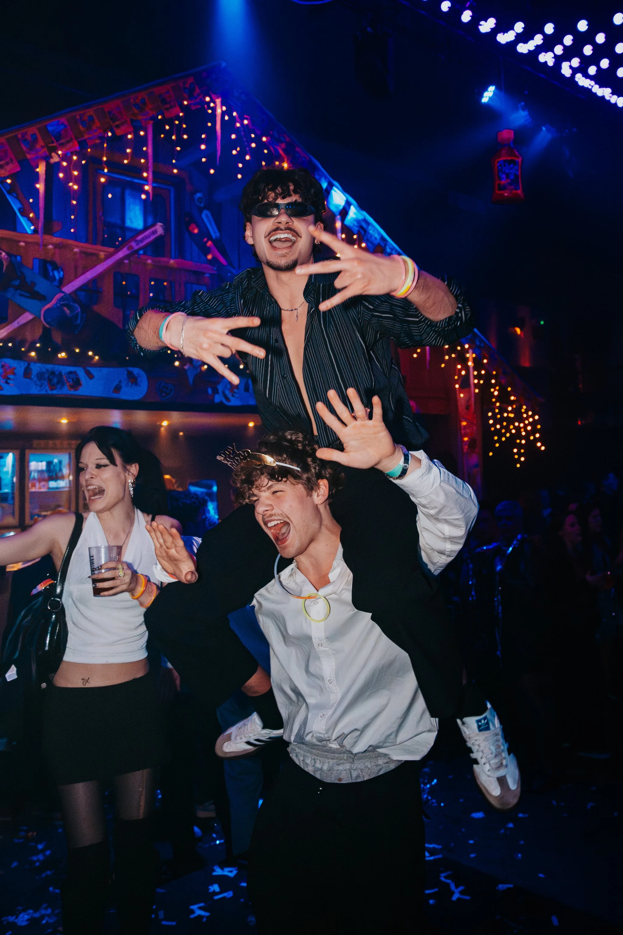 People celebrating at a party with colorful lights, confetti on the floor, and decorations hanging from the ceiling. A person in sunglasses is being held up, smiling, while others dance around.