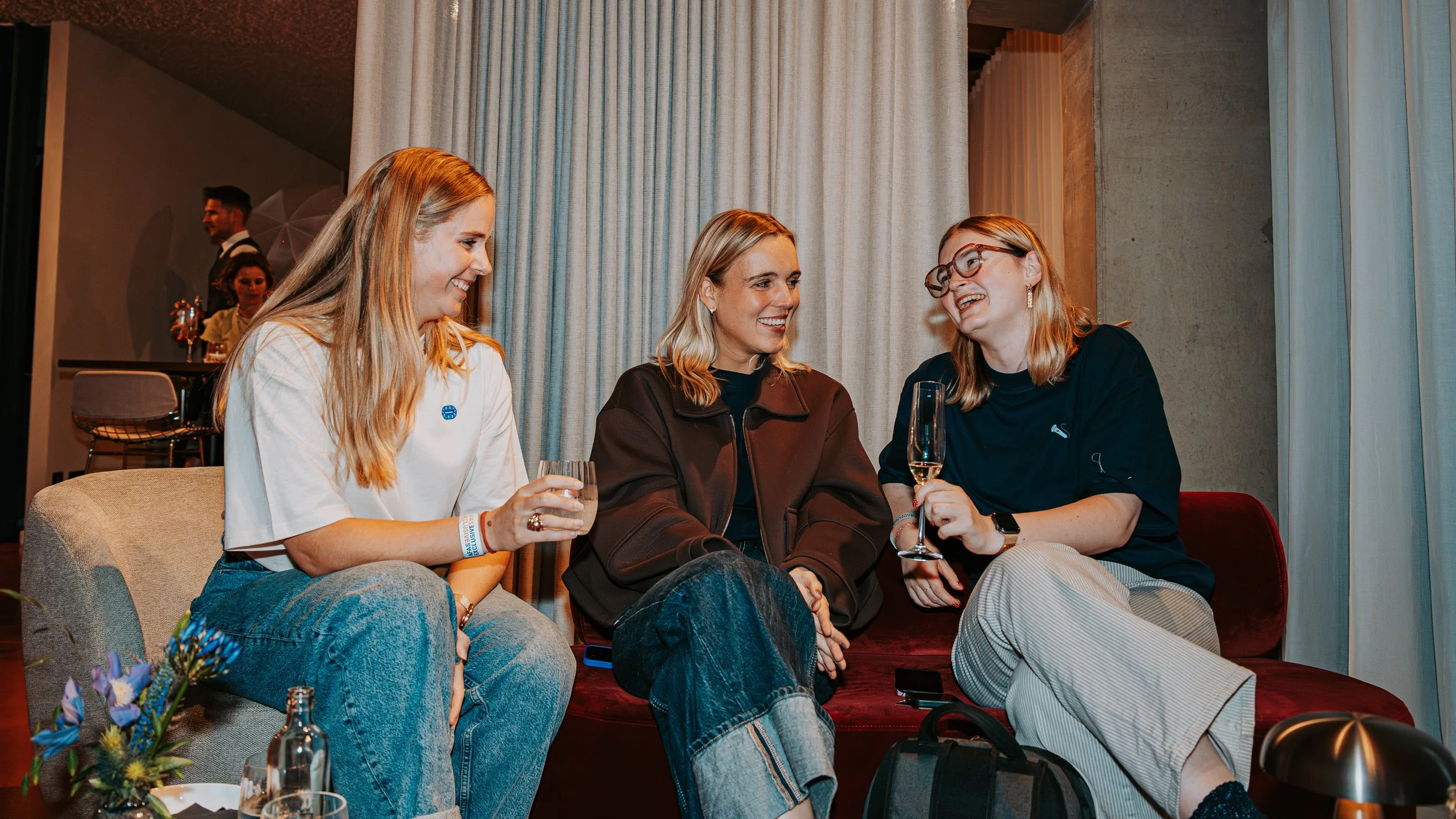 Three women sitting on a sofa and engaging in conversation while holding champagne glasses at a social event. They appear happy and are smiling.