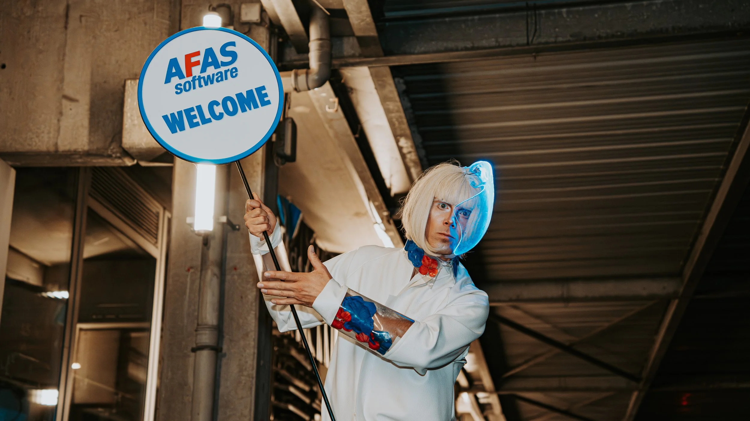 Person dressed as a scientist with light blond hair, white lab coat with blue details, and a translucent visor, holding a round sign that says 'AFAS software WELCOME' in a setting with industrial ceiling and fluorescent lighting.