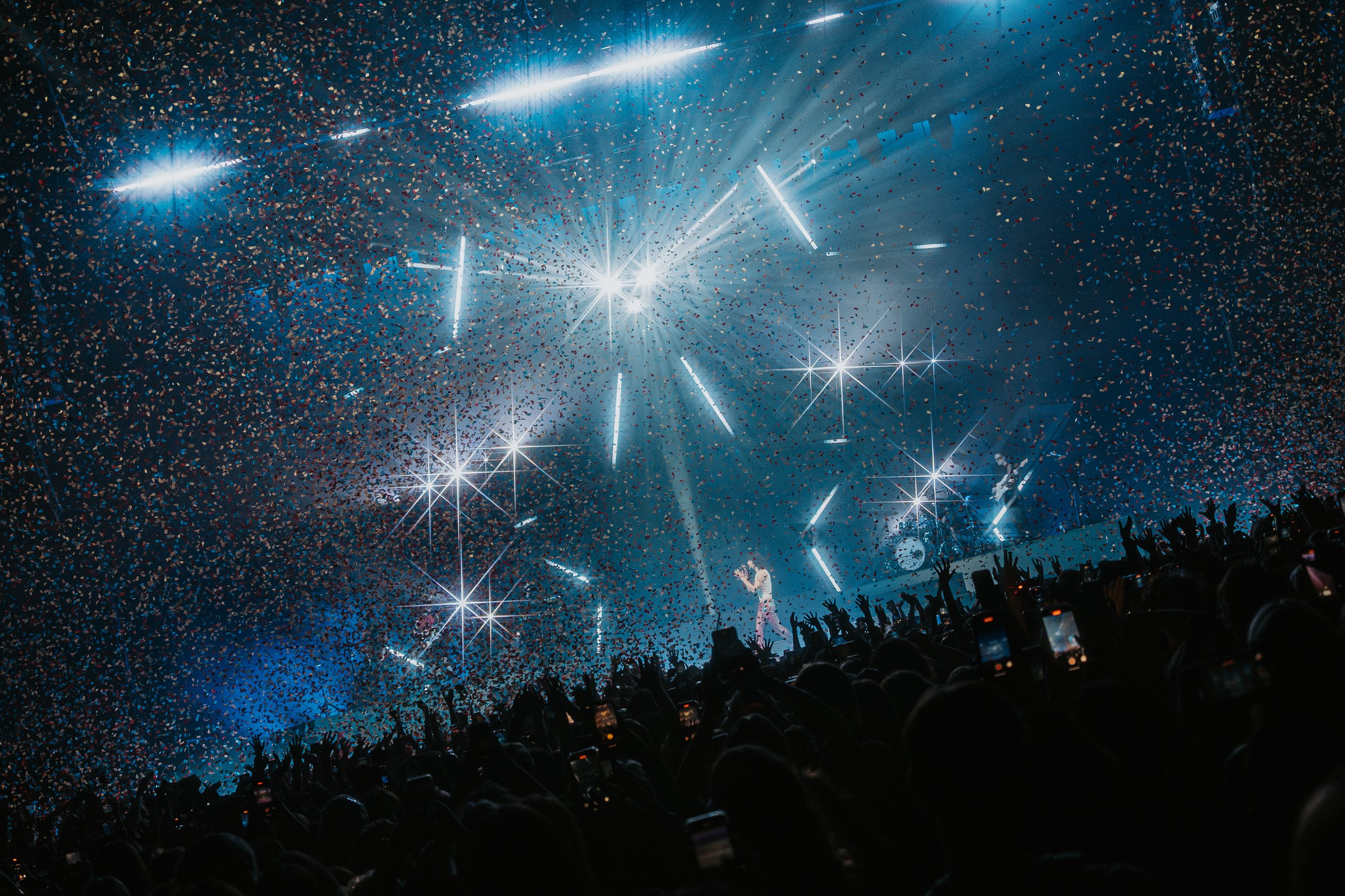 A concert scene with bright stage lights, colorful confetti, and silhouettes of cheering audience members with their hands raised.