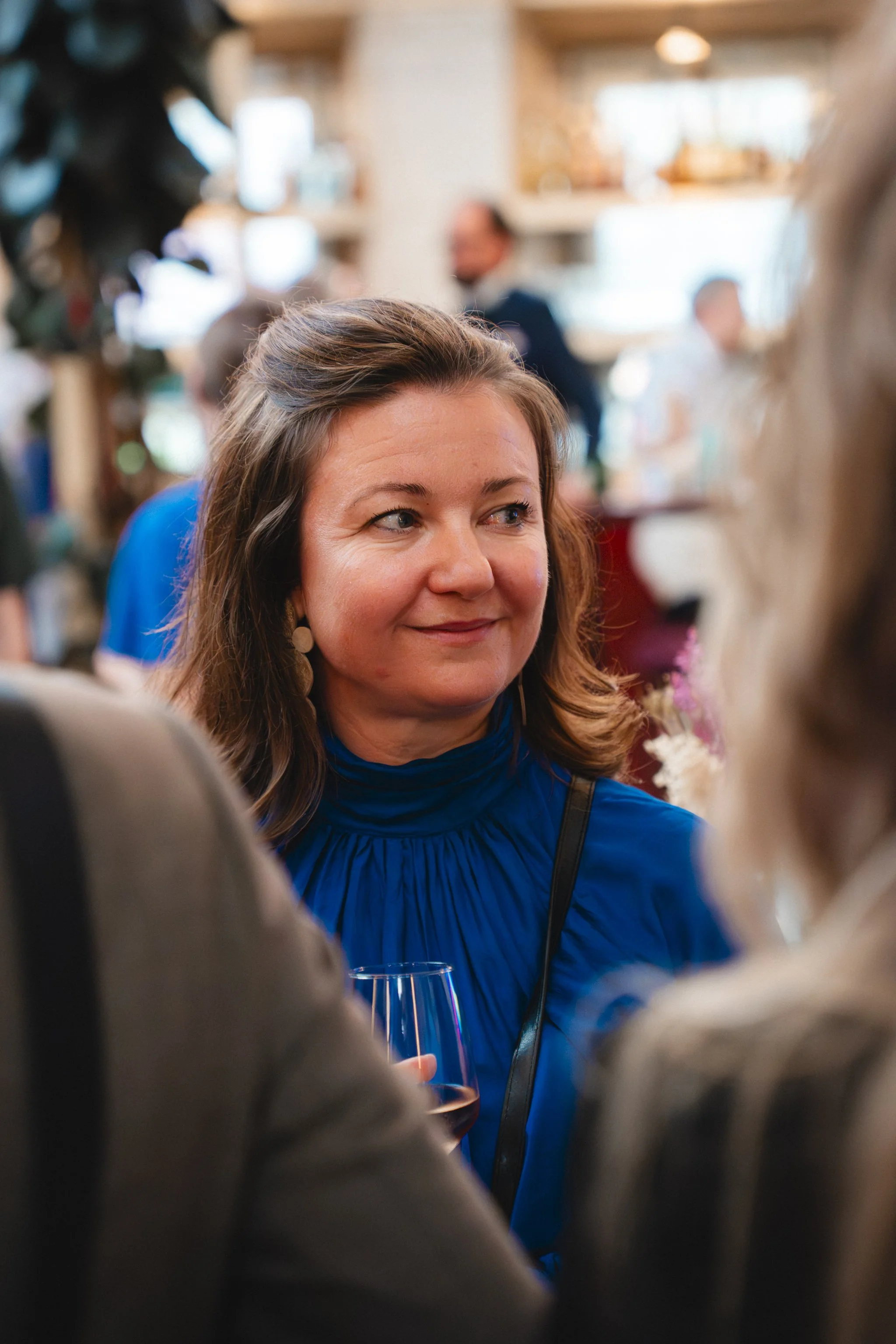 A woman with light skin, brown hair, and wearing a blue jacket is smiling in a crowded indoor setting. She is holding a glass of wine and looking to her left. The background is blurred, showing other people and a well-lit interior.