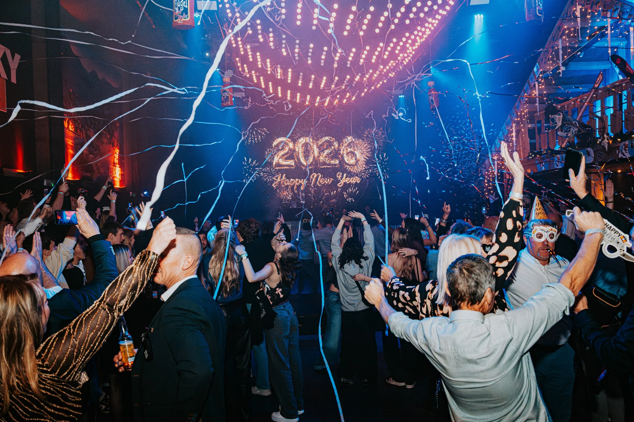 Crowd of people celebrating New Year's Eve with confetti, fireworks, and a large illuminated '2020' sign in the background.