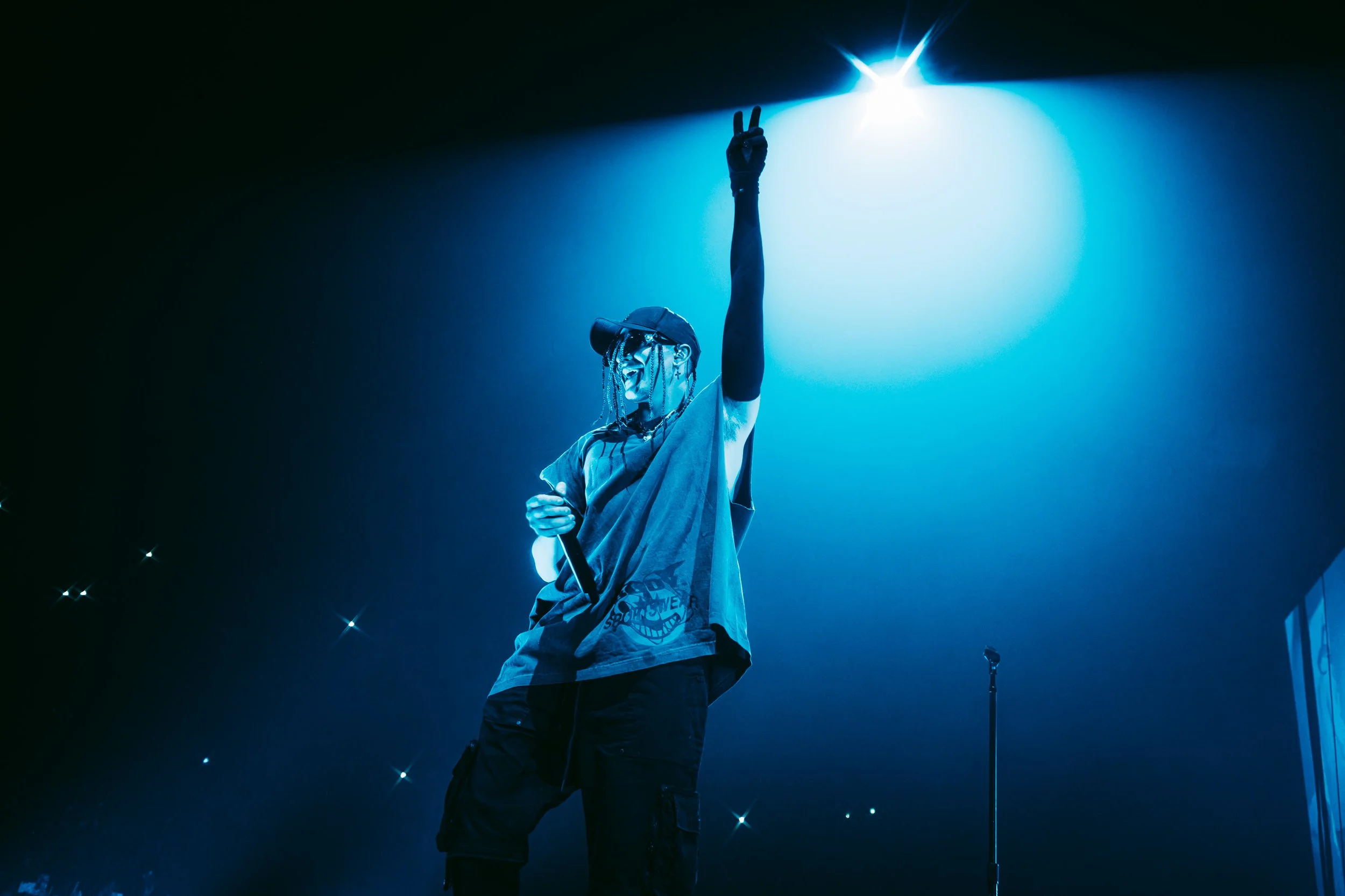 Person performing on stage under blue spotlight, holding a microphone, wearing a cap, glasses, and casual clothing, making a peace sign with their hand.