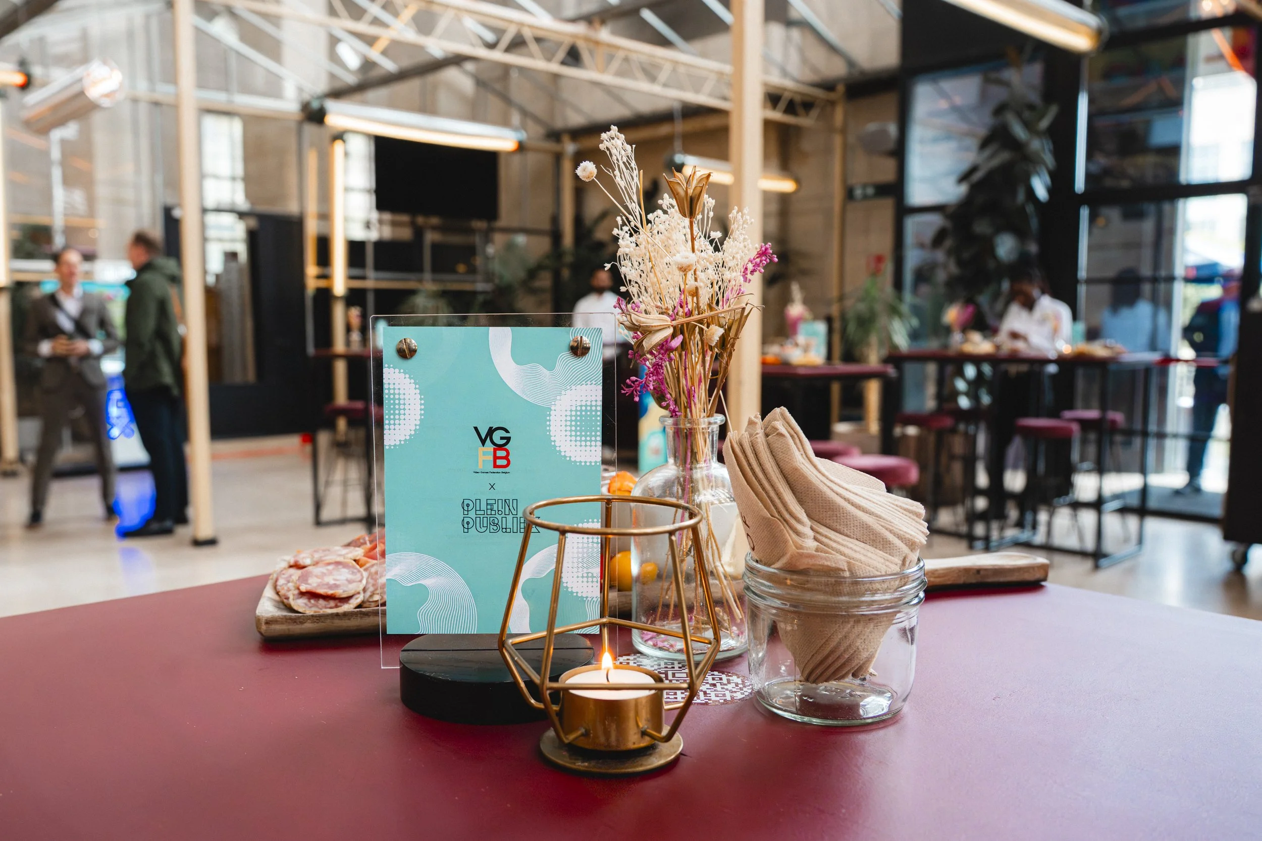 A table display with dried flower arrangements, a glass jar with folded napkins, a lit candle, a small sign with a blue design, and some slices of pizza. The background shows a modern, industrial-style cafe with people and wooden and metal decor.
